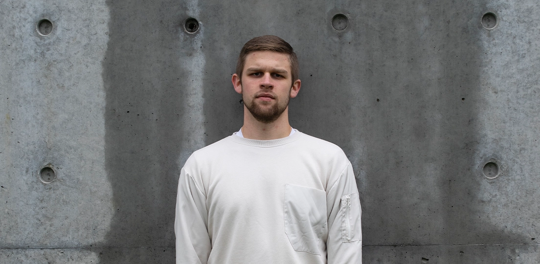 Stark image of a young man with short hair and a beard wearing a white sweatshirt standing with a serious expression against a textured concrete wall.