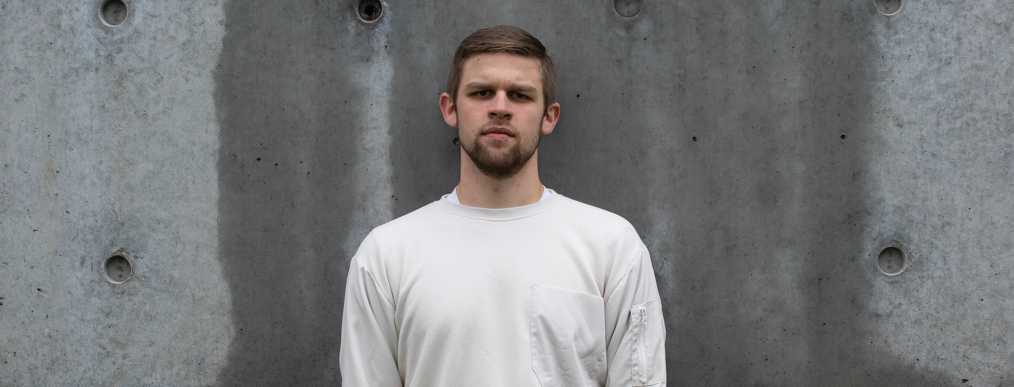 Stark image of a young man with short hair and a beard wearing a white sweatshirt standing with a serious expression against a textured concrete wall.