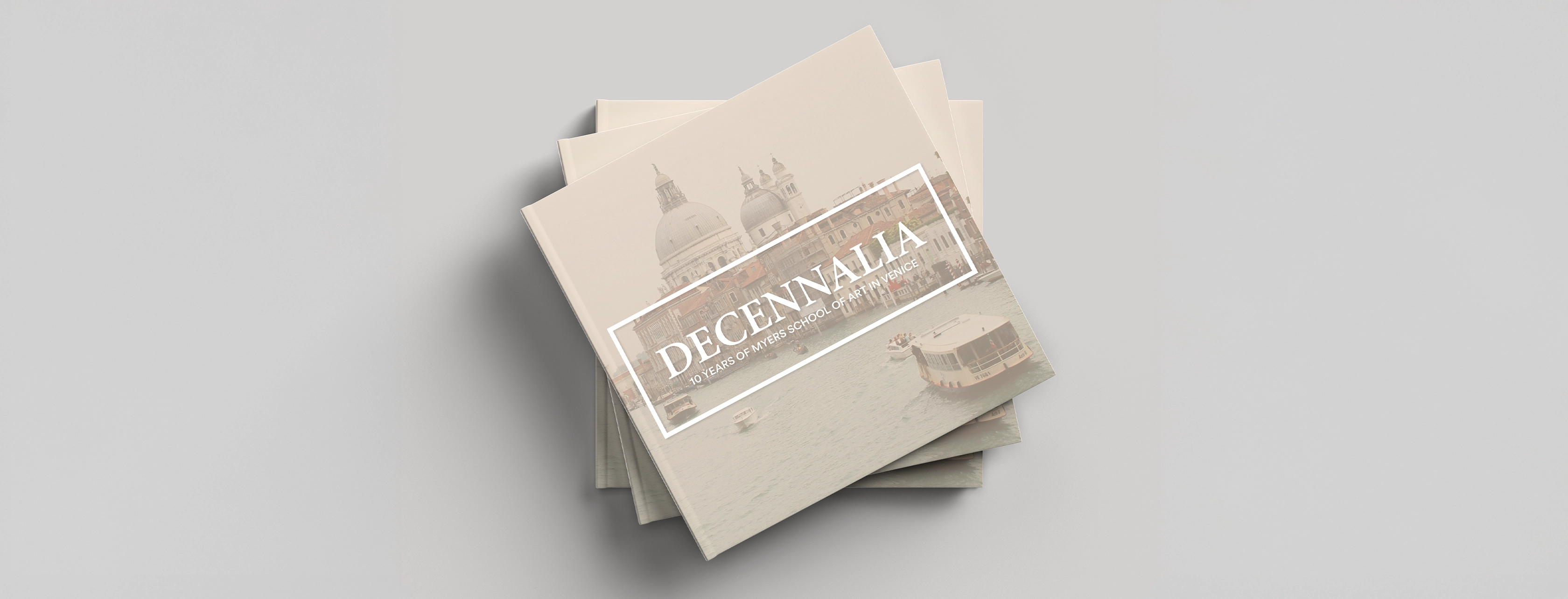 Stack of tan books titled 'DECENNALIA 10 Years of Myers School of Art in Venice' with cover image of Venice buildings and boats on water.