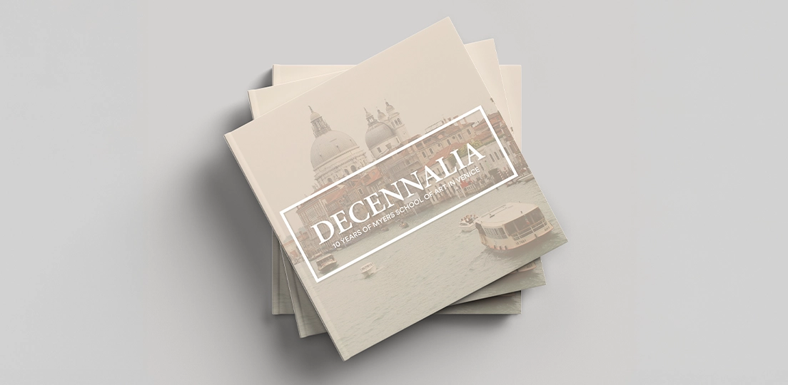 Stack of tan books titled 'DECENNALIA 10 Years of Myers School of Art in Venice' with cover image of Venice buildings and boats on water.
