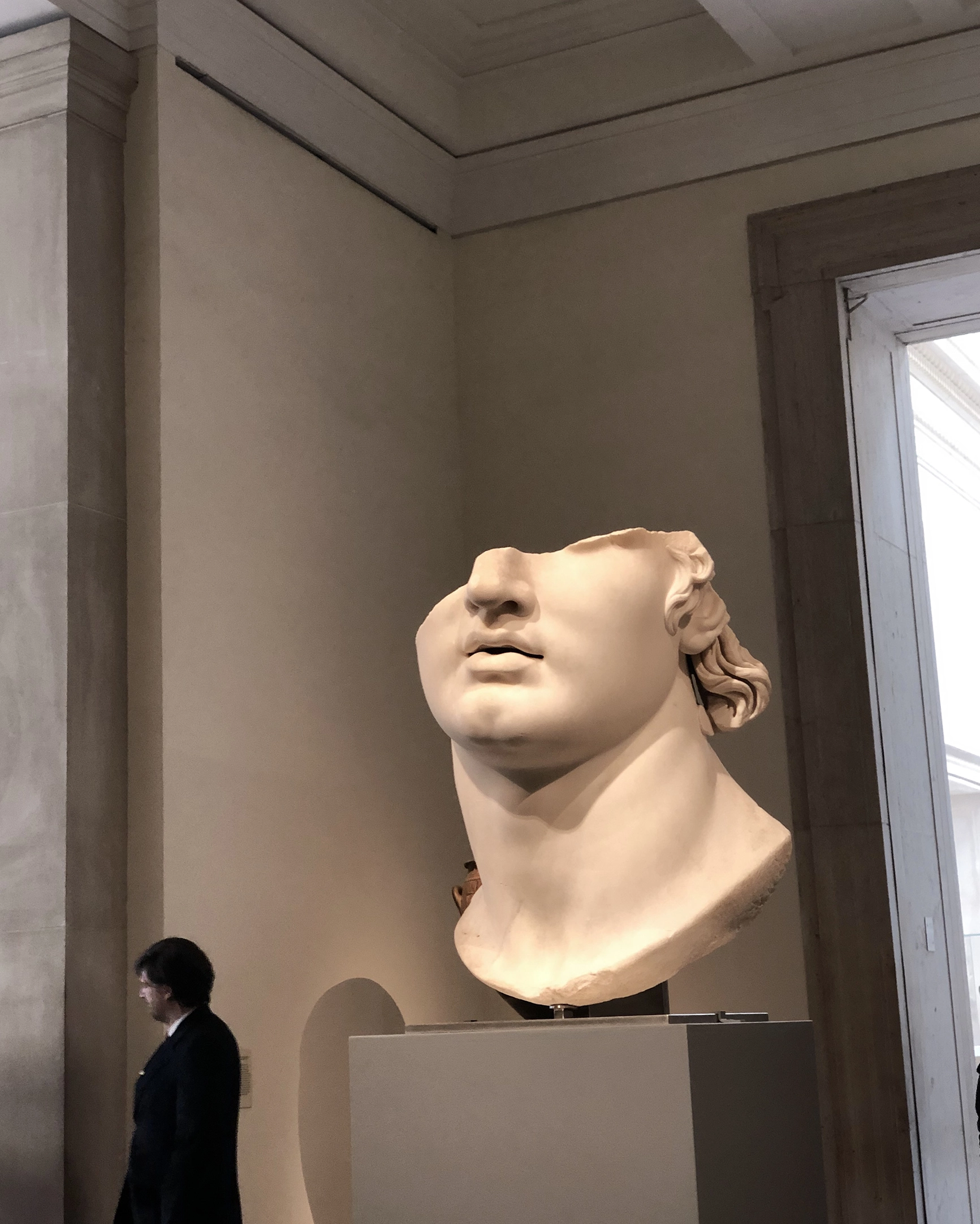 Large classical marble sculpture of a head missing the upper part, displayed on a pedestal in a museum.