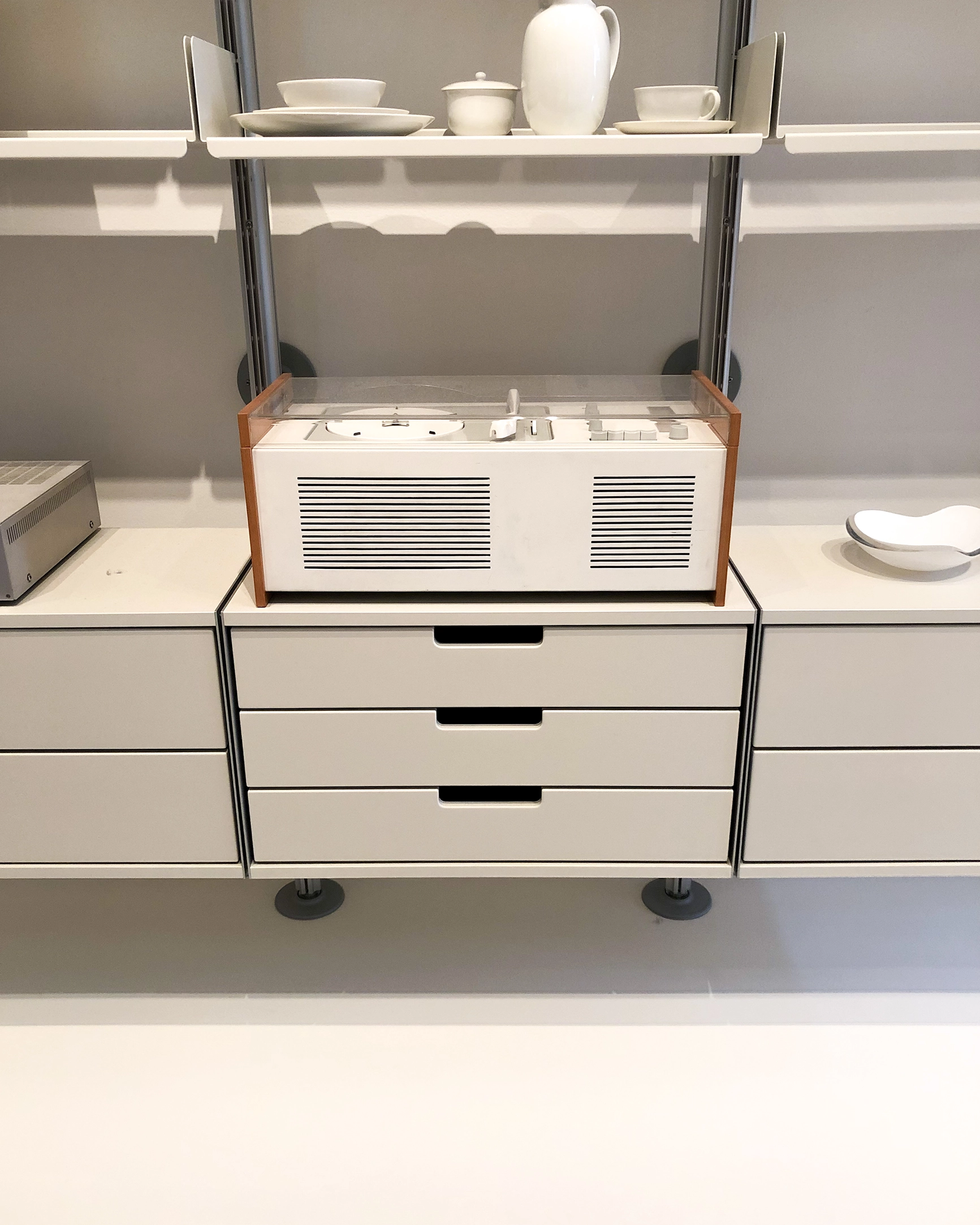 White minimalist stereo system with wood sides on modern white wall-mounted drawers under a shelf holding white ceramic dishes.