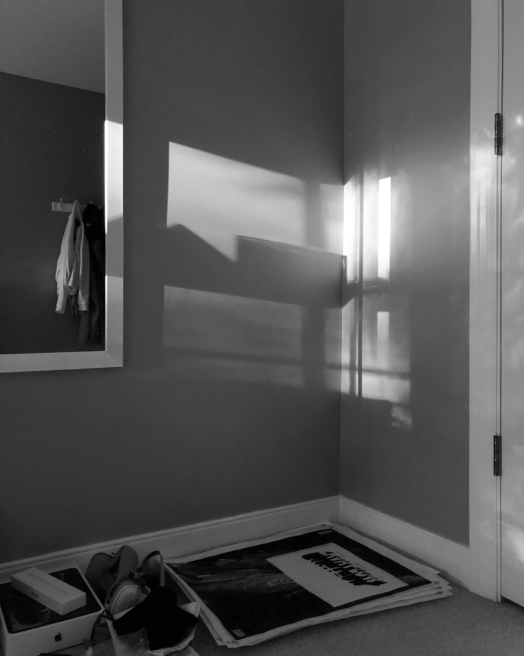 Black and white photo of the corner of a room with sunlight casting window shadows on gray walls, a white door, and some items on the carpeted floor.
