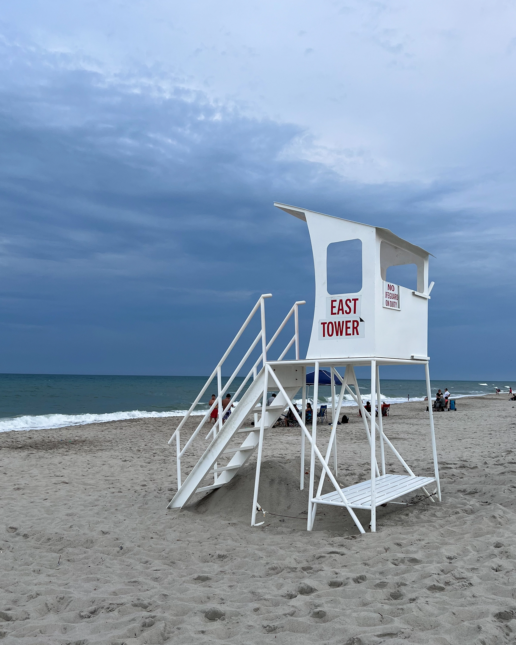 White lifeguard tower labeled East Tower on a sandy beach under a cloudy sky with ocean waves in the background.