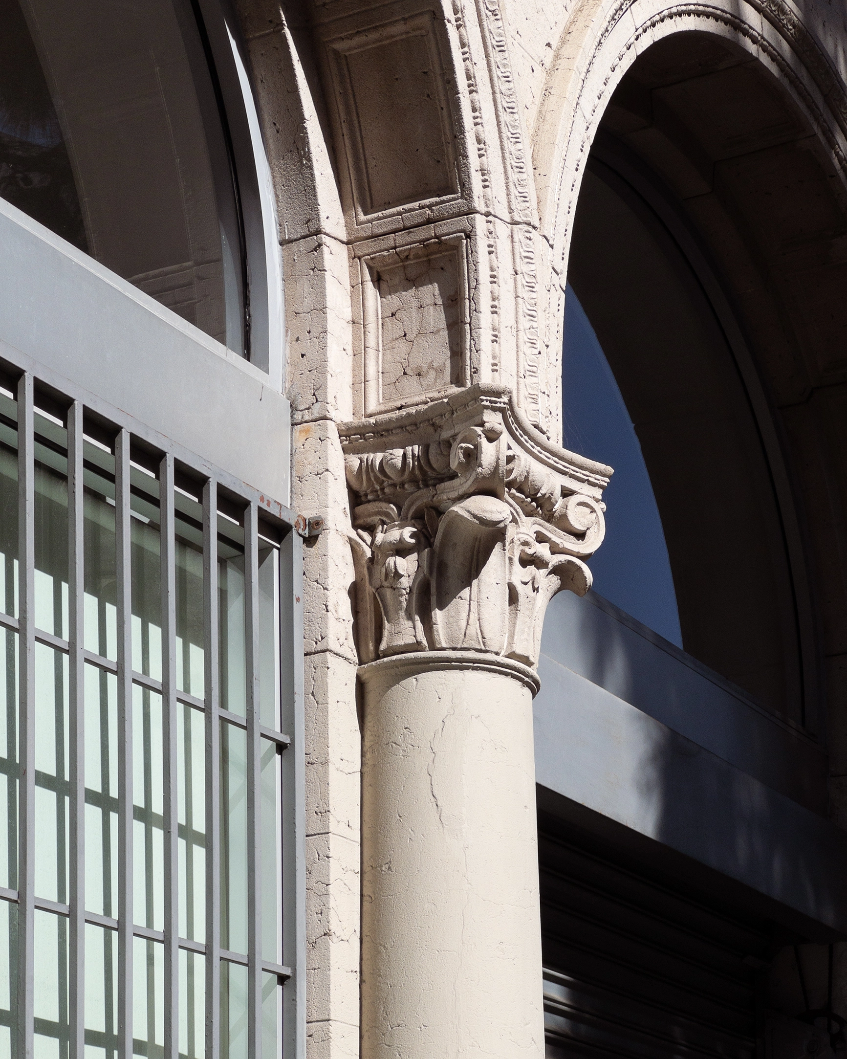 Close-up of an ornate beige stone column with a carved Corinthian capital next to a tall light blue window with light and thin metal bars.