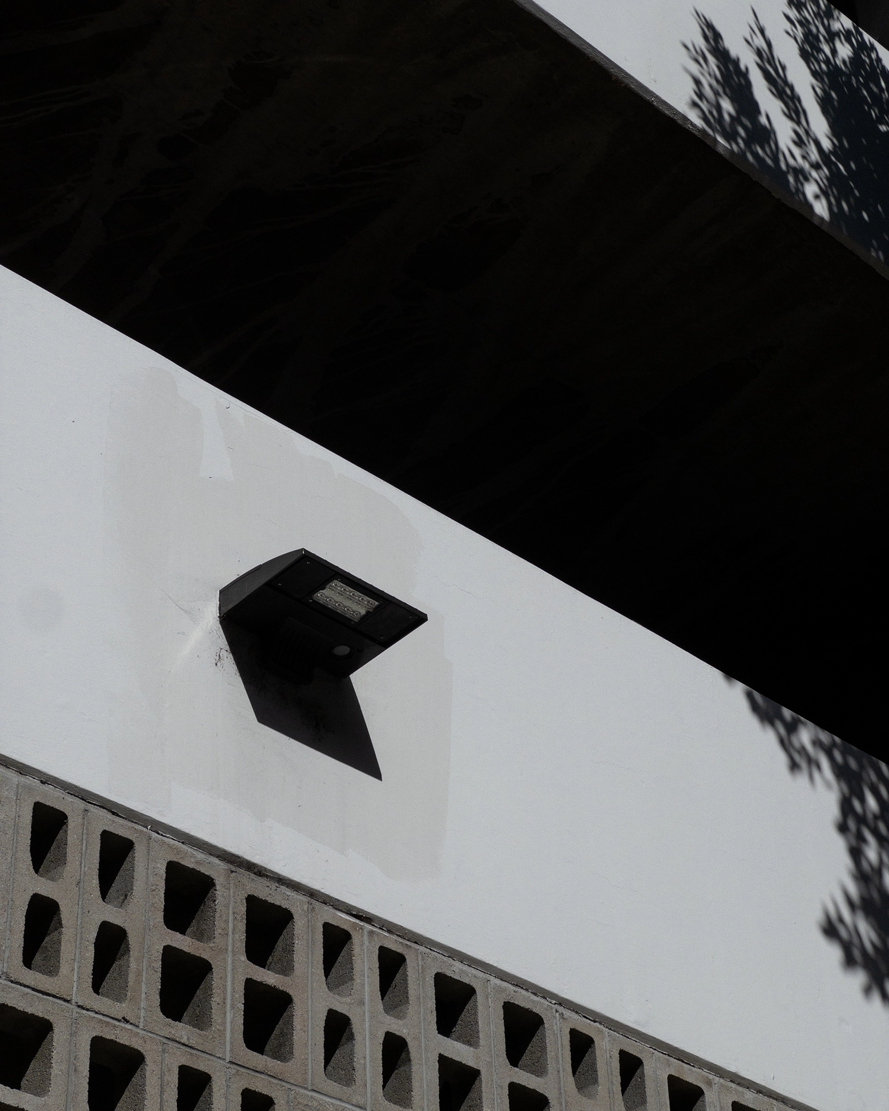 Modern black wall-mounted outdoor light fixture casting a stark rectangular shadow on a gray wall above concrete block ventilation openings.