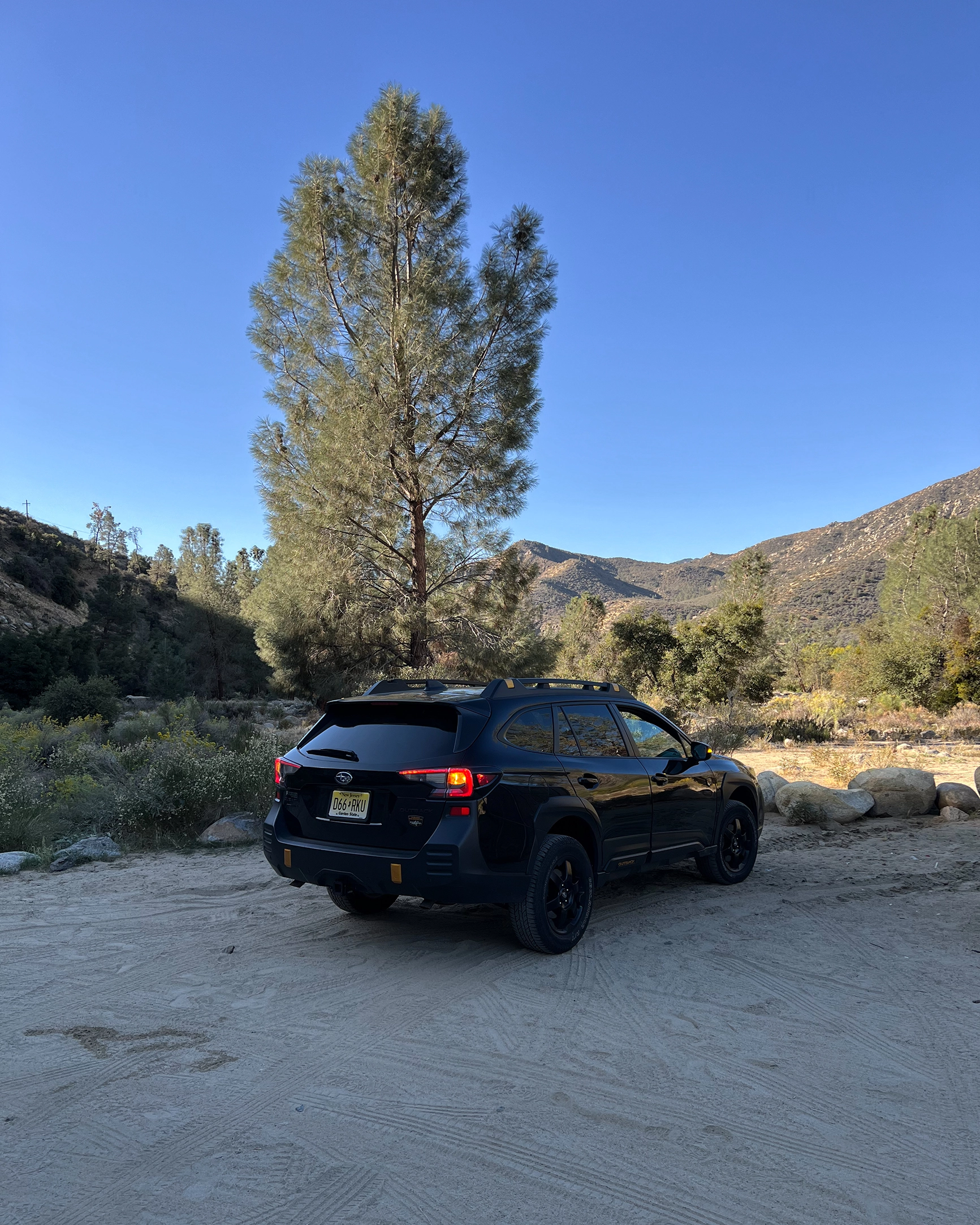 Black Subaru Outback parked on a desert area near trees and rocky hills under a clear blue sky.