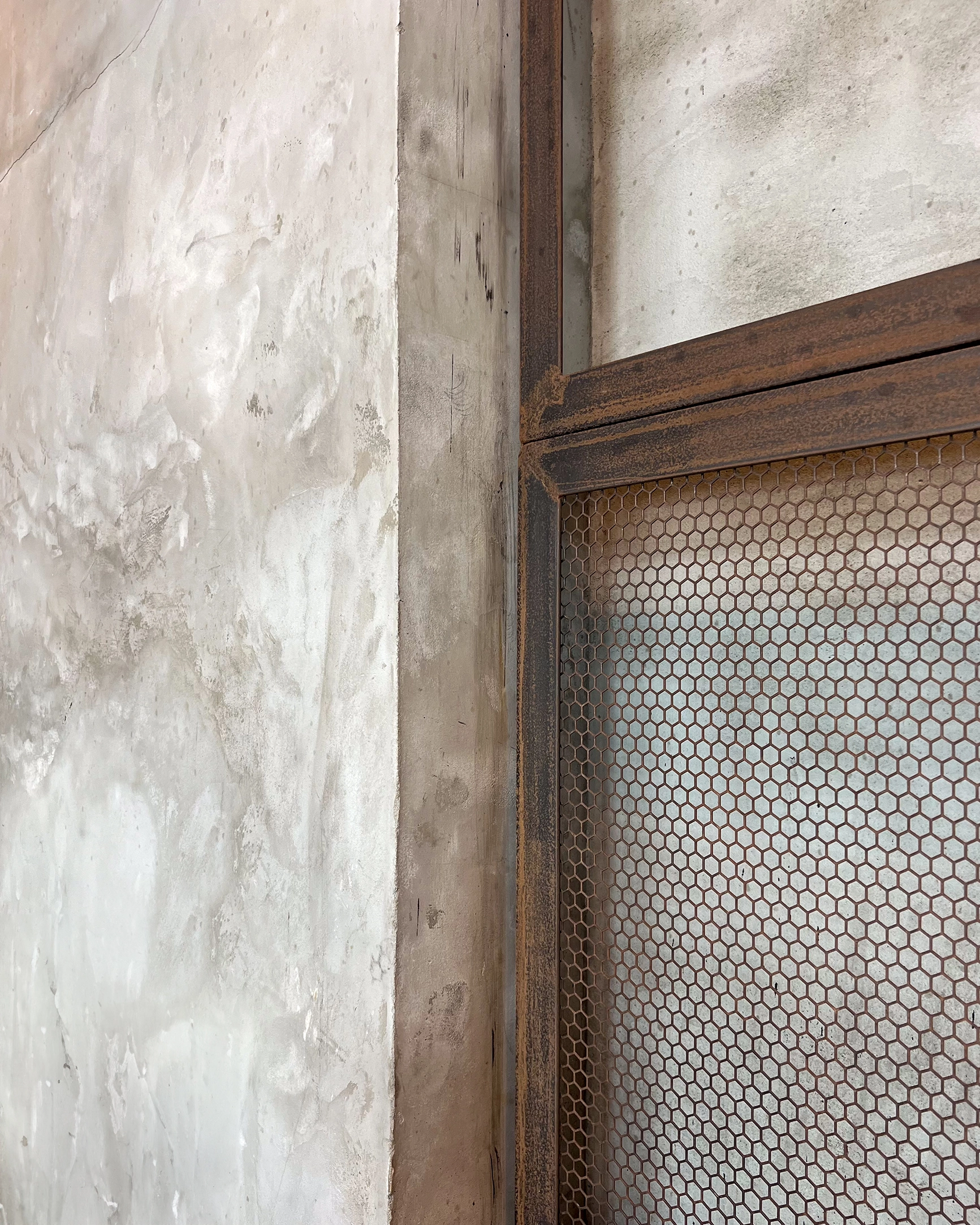 Close-up of a textured concrete wall next to a rusted metal mesh window frame.