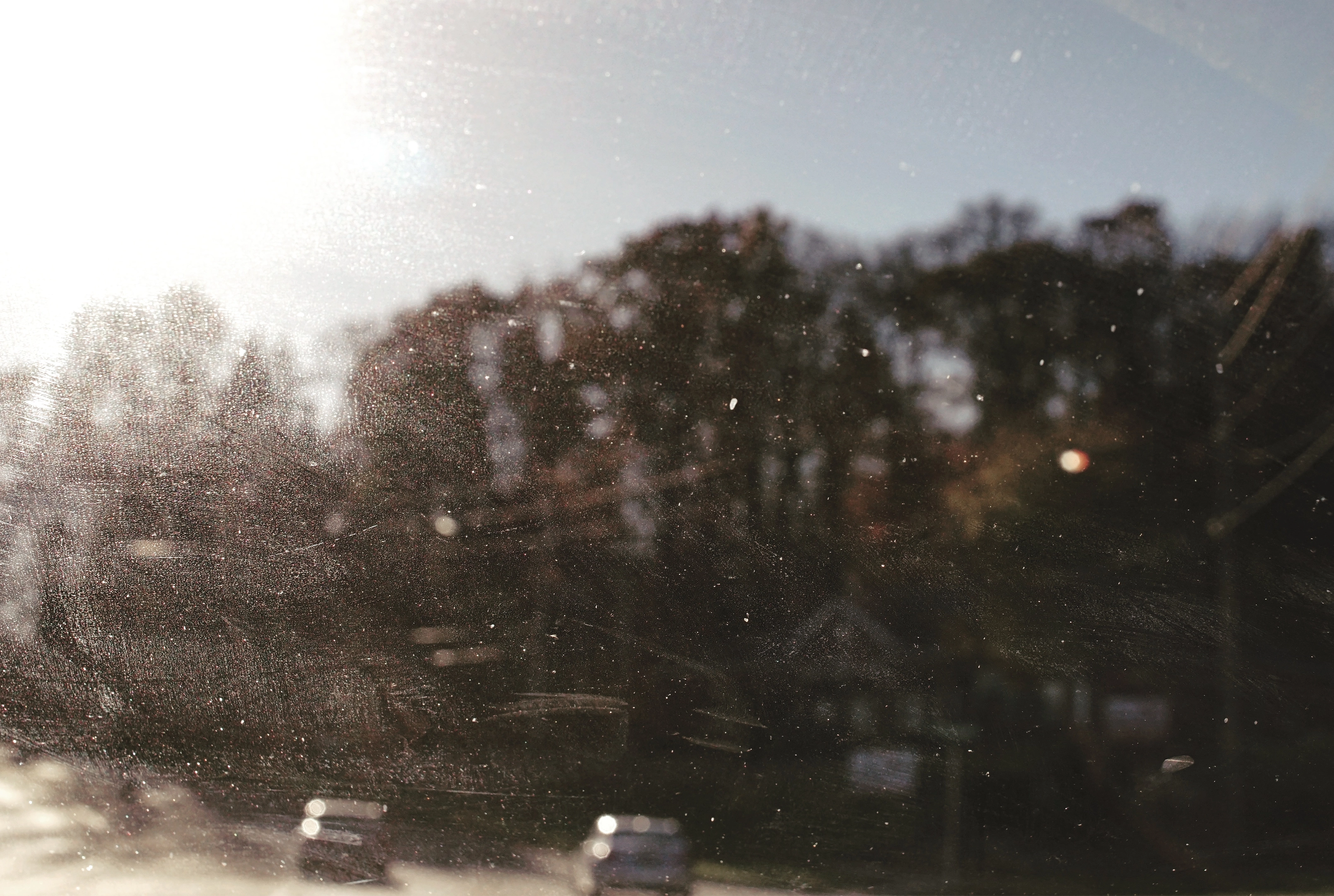 Blurry view through a dirty window showing cars on a road and orangey trees in the background under bright sunlight.