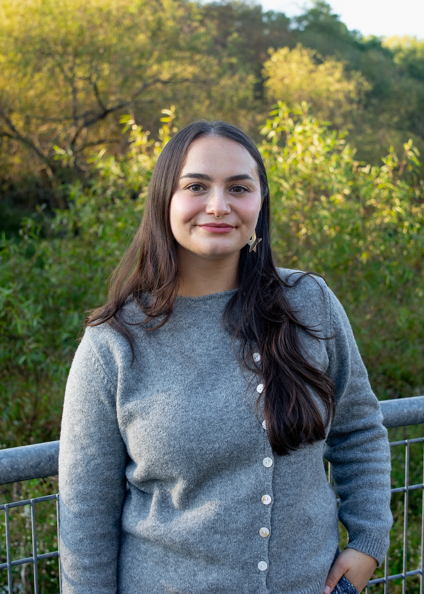 Young woman with long dark hair wearing a grey cardigan and star-shaped earrings standing outdoors near a metal railing with light green and yellow trees in the background.