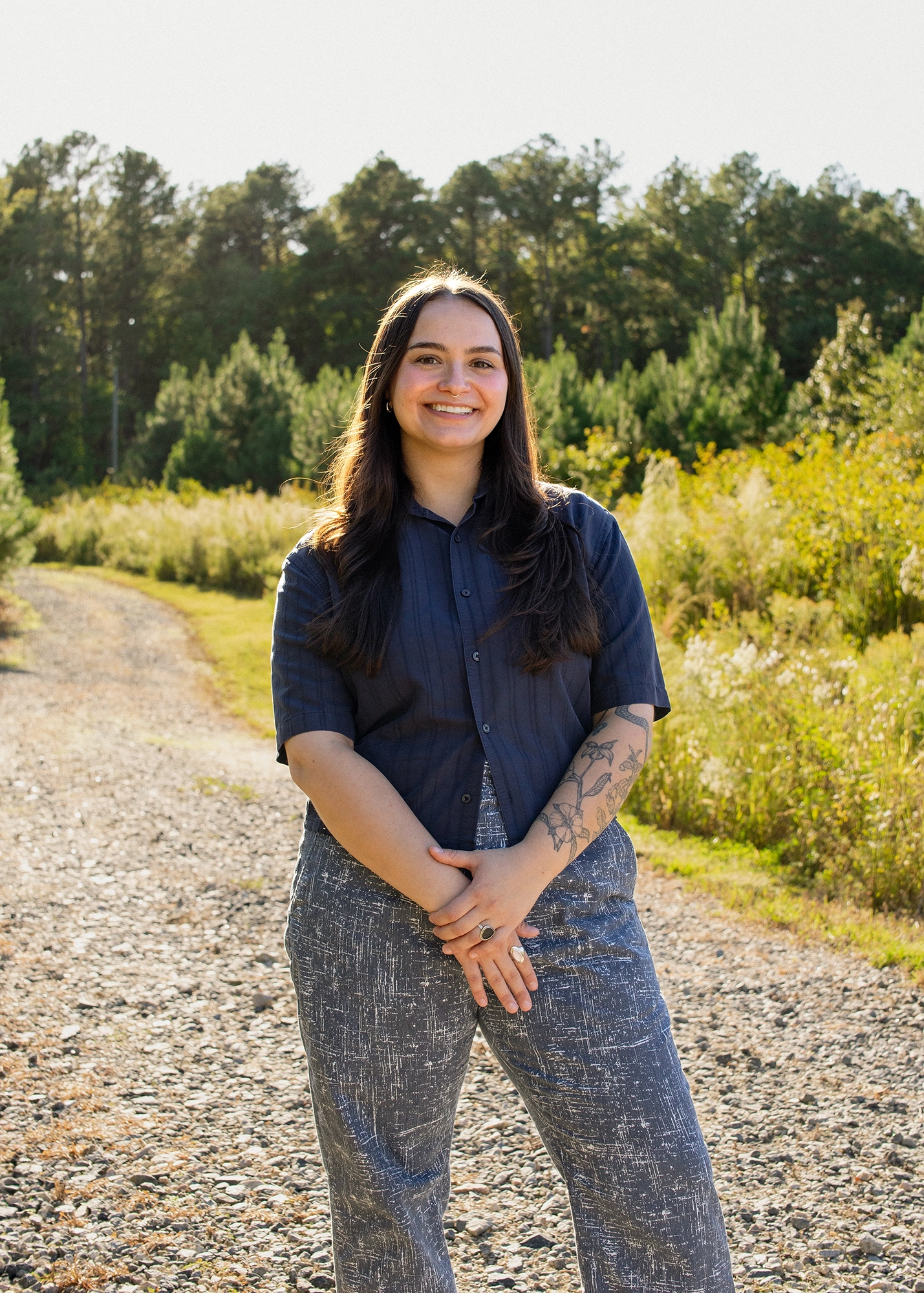 Young woman with long dark hair and floral arm tattoo smiling, and standing on a whimsical sunlit gravel path surrounded by yellow tall grass and green pine trees.