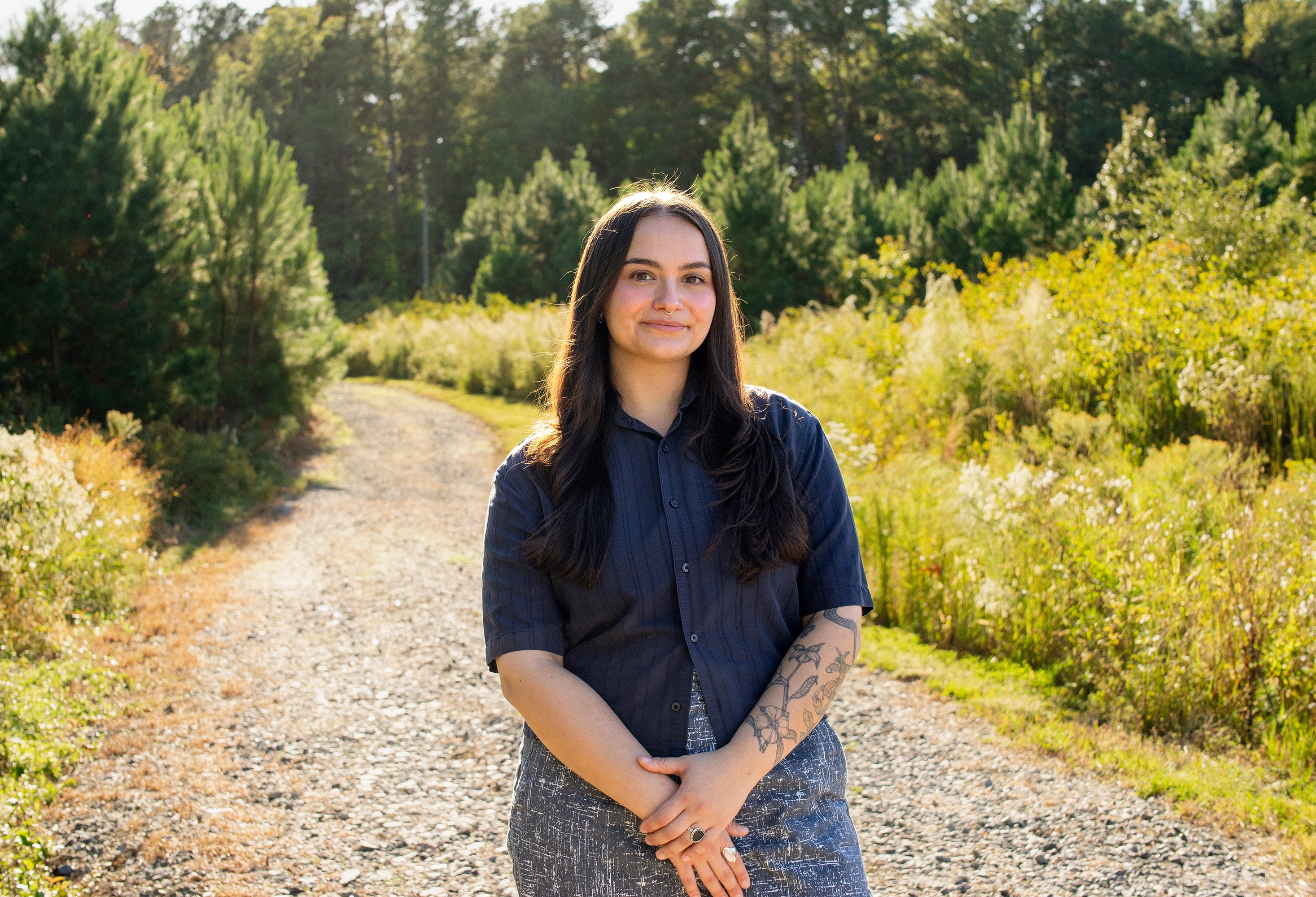 Young woman with long dark hair and floral arm tattoo standing on a whimsical sunlit gravel path surrounded by yellow tall grass and green pine trees.