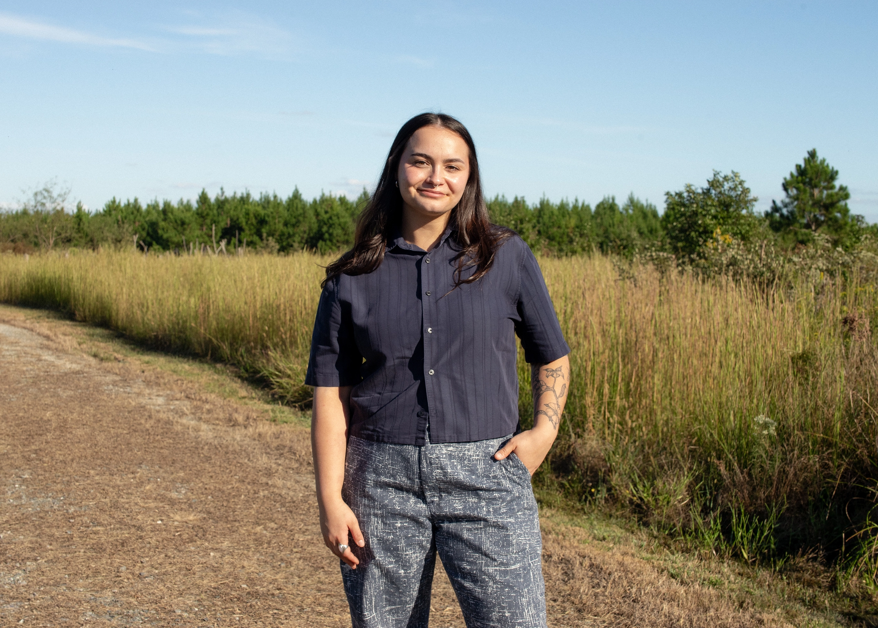 Young woman with long dark hair and a slight smile, wearing a navy short-sleeve shirt and patterned gray pants, standing on a dirt path next to tall grass under a clear blue sky.