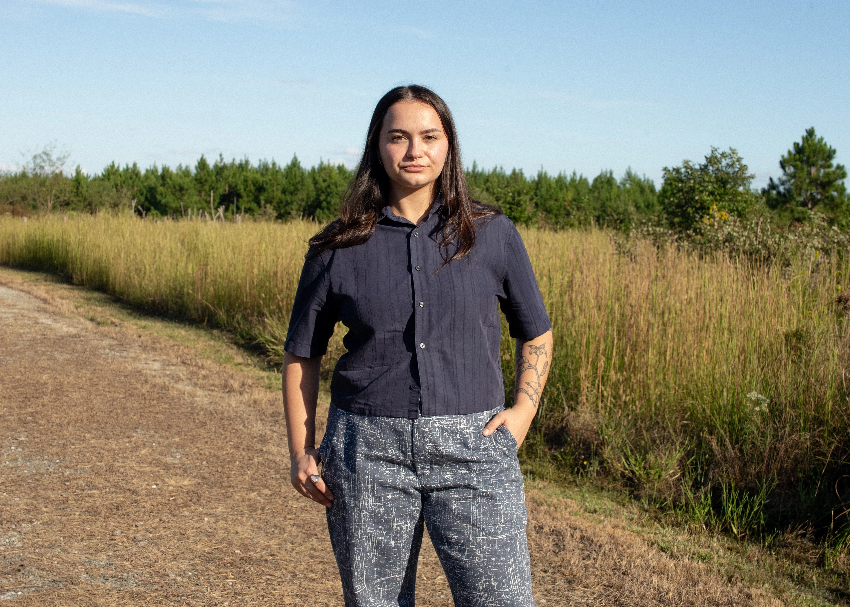 Young woman with long dark hair and a serious expression, wearing a navy short-sleeve shirt and patterned gray pants, standing on a dirt path next to tall grass under a clear blue sky.
