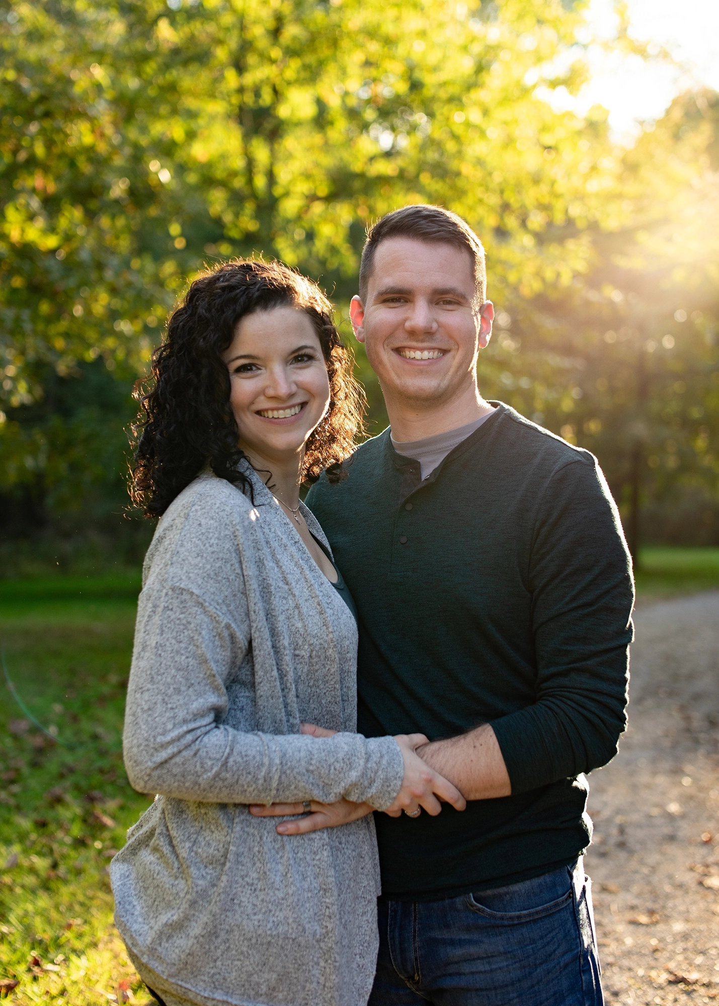 Smiling couple standing outdoors in sunlight with green trees and pathway in the background.