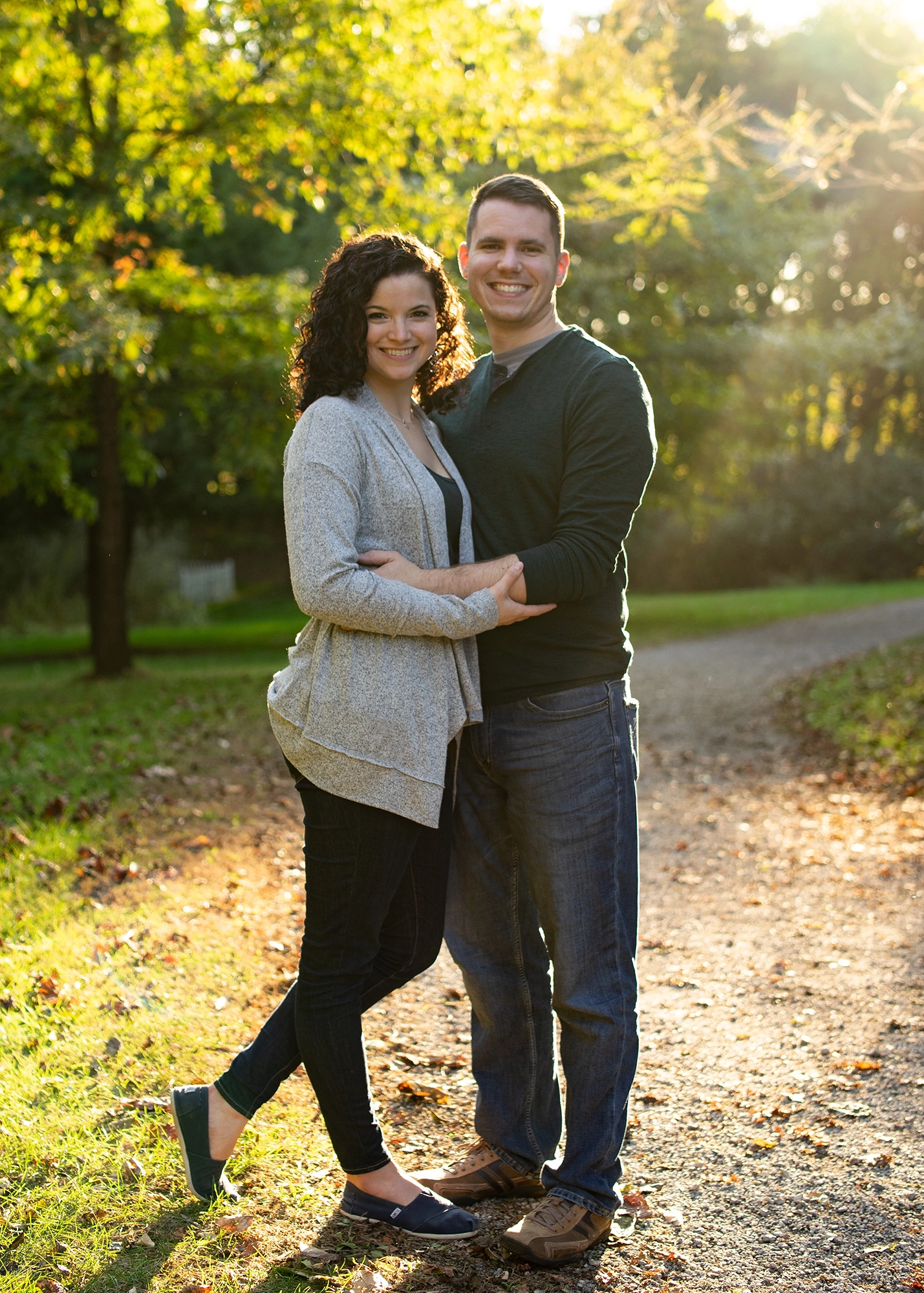 Smiling couple embracing on a sunlit wooded path with green trees in the background.