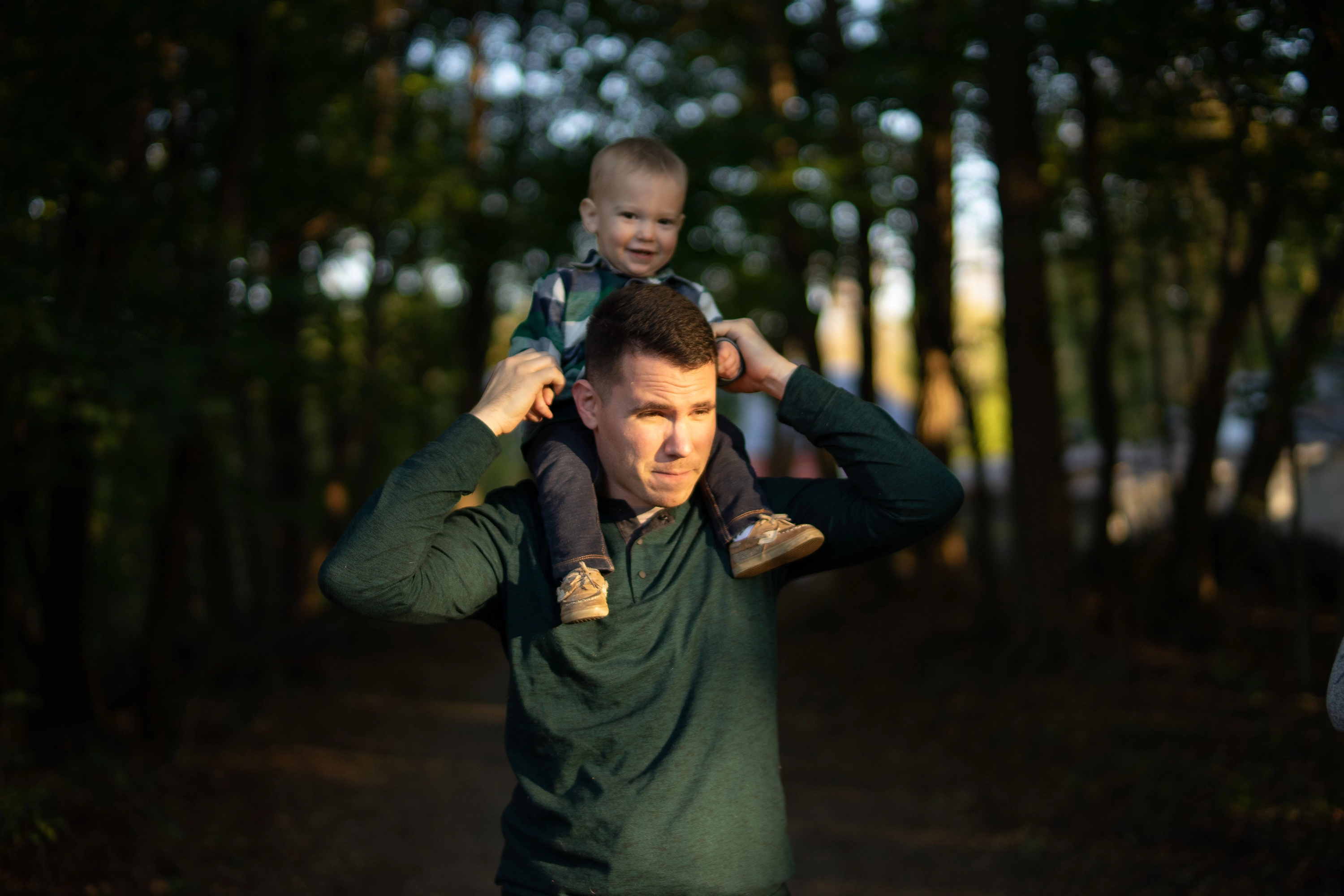 Father wearing a dark green shirt and with golden sunlight on his face carrying a smiling toddler on his shoulders in a wooded area.