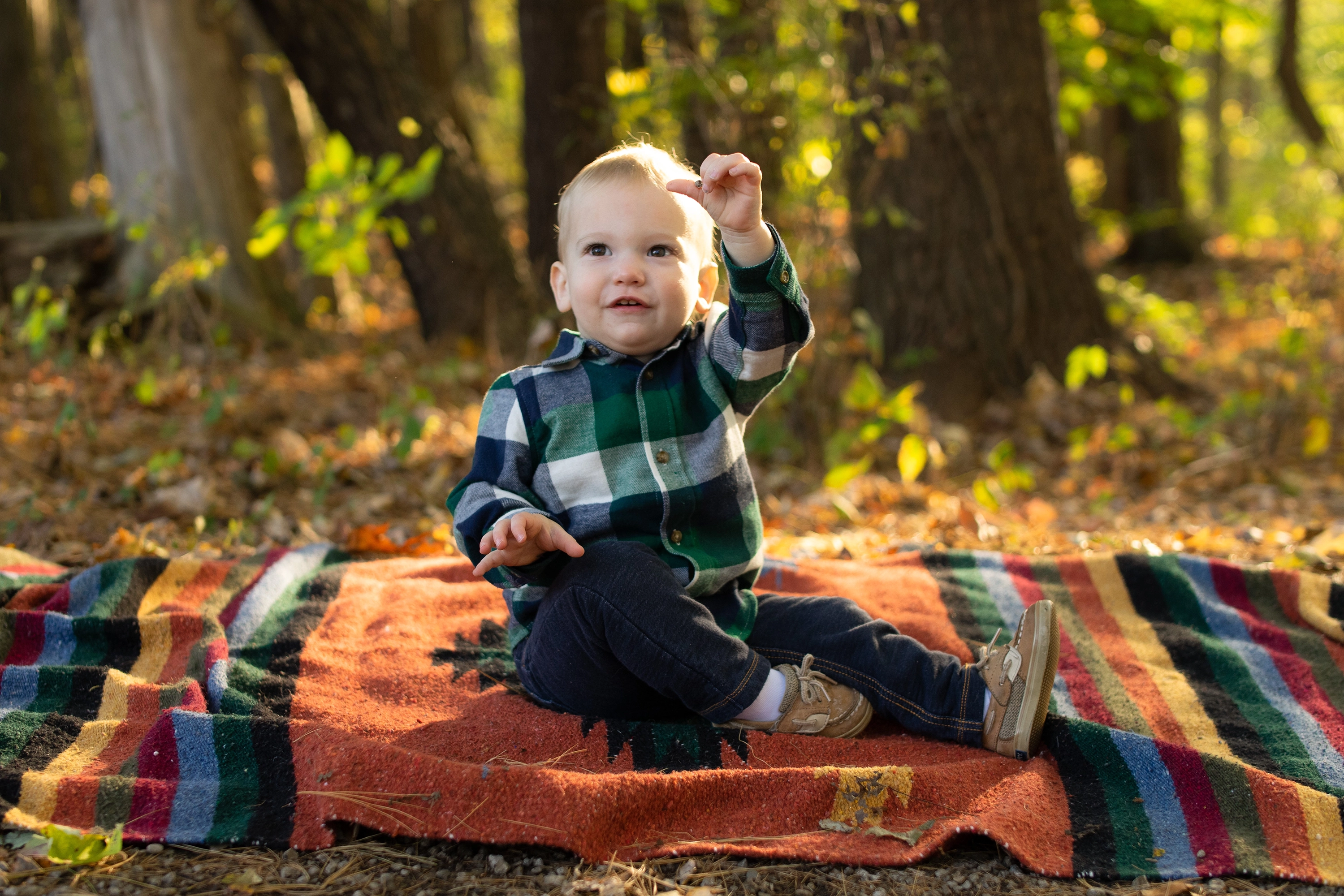 Toddler sitting on a colorful striped blanket in a forest, holding up something and looking slightly up.