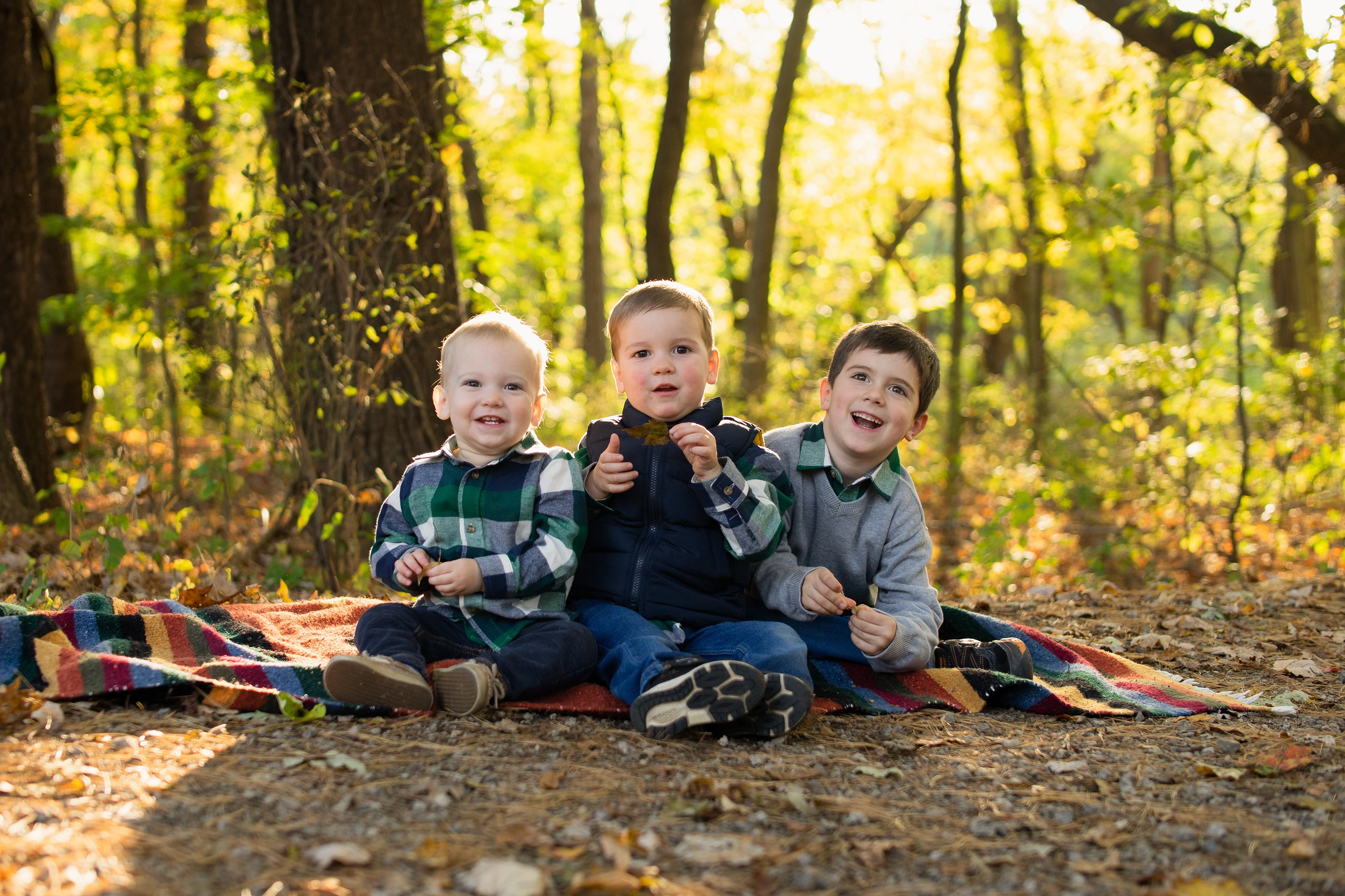 Three smiling young boys sitting on a colorful blanket in a sunlit forest during autumn.