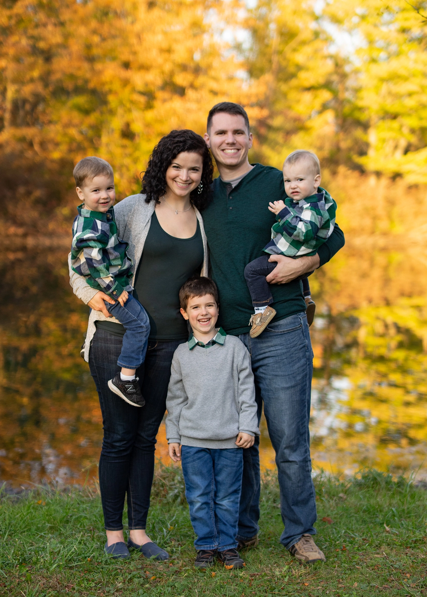 Smiling family of five with two parents holding toddlers and one child standing between them outdoors with vibrant yellow autumn trees in background.