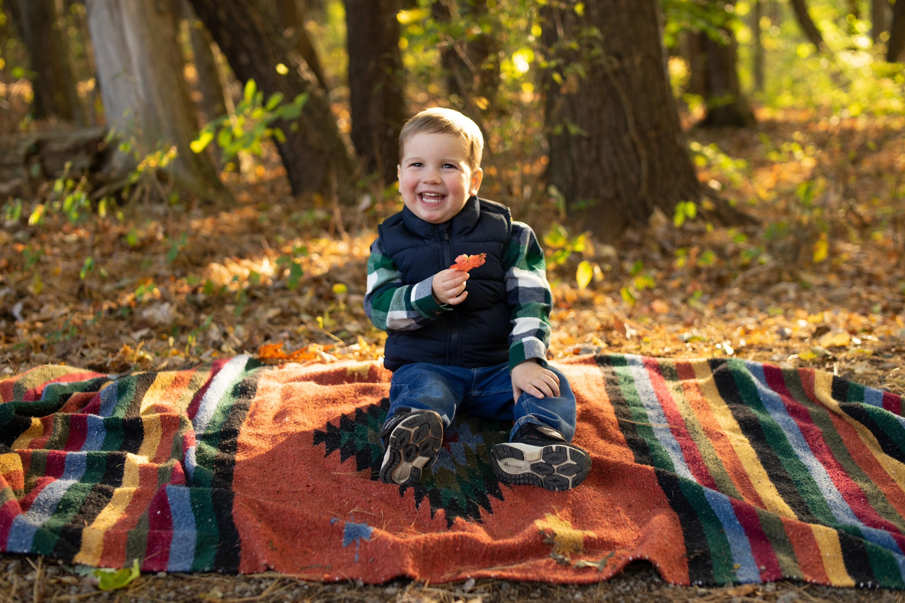 Smiling toddler sitting on a colorful striped blanket outdoors in a forest holding an autumn leaf.