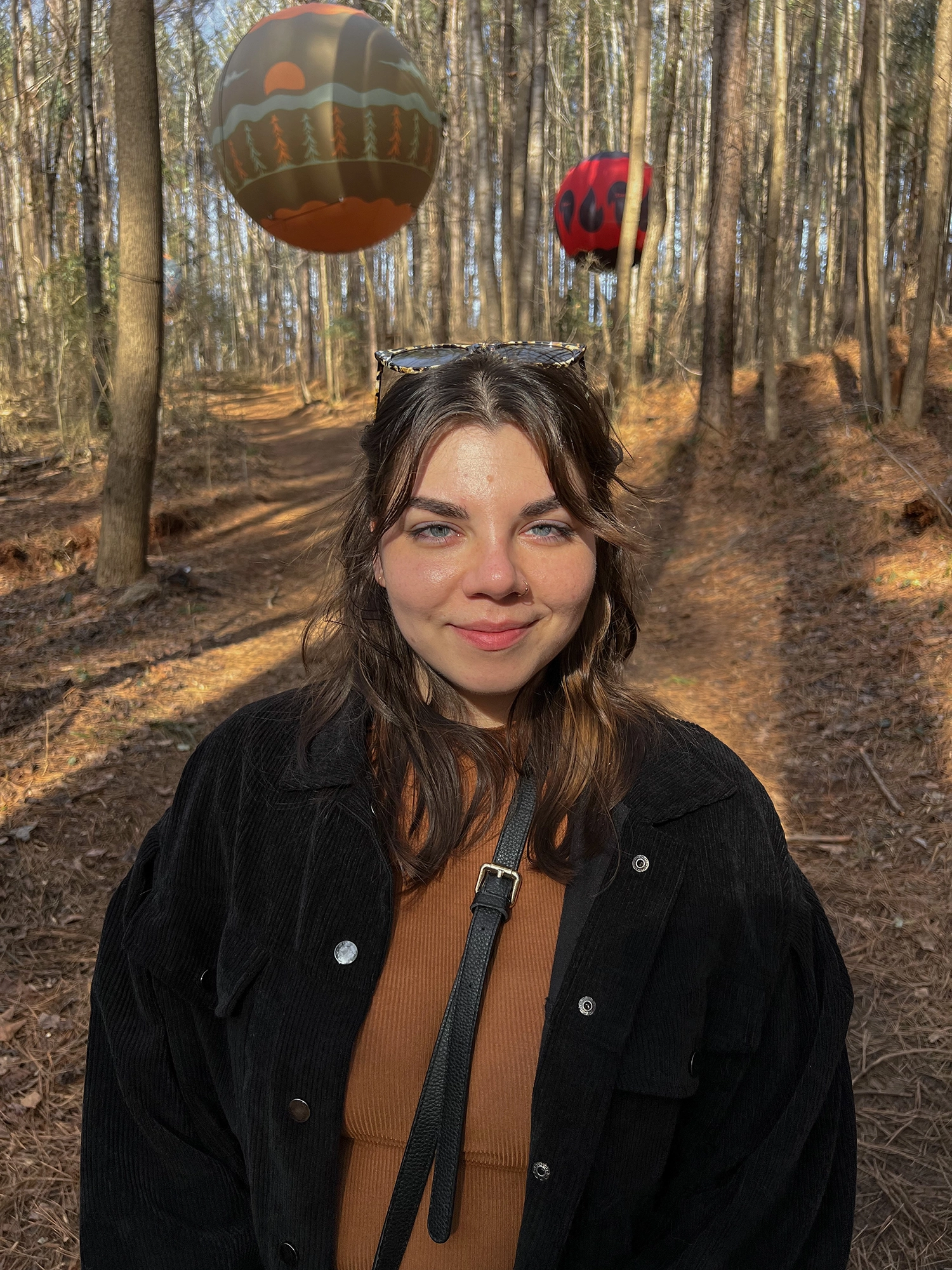 Portrait photo of a young woman with brown hair and blue eyes standing on a barren but warm forest path with two decorative balloons floating behind her.