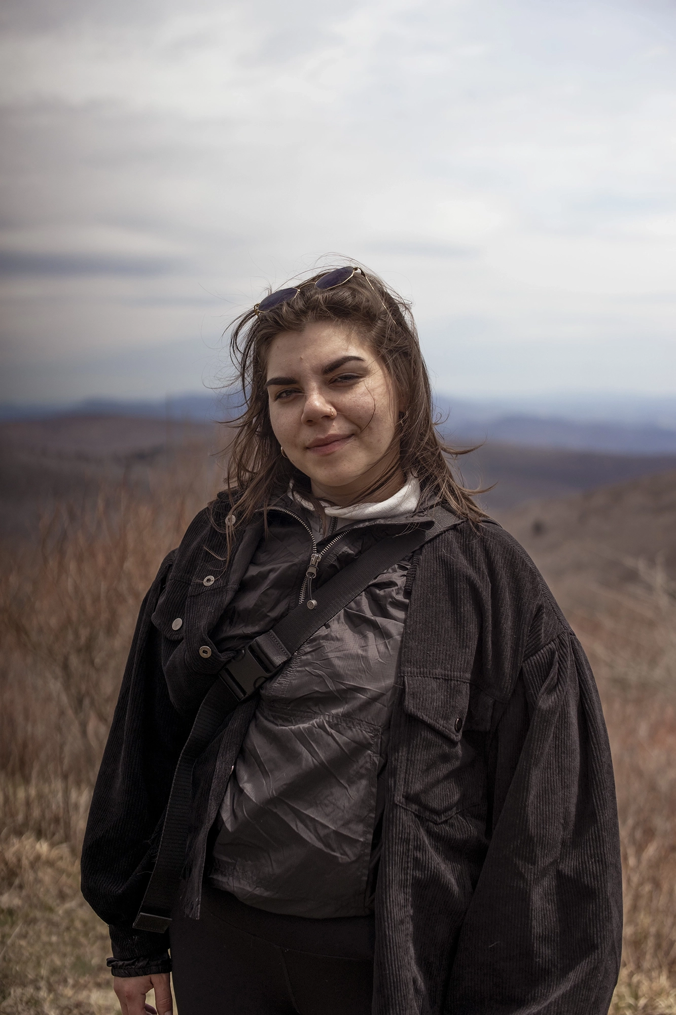 Young woman with brown hair wearing a black jacket standing outdoors with a reddish-blue mountain landscape in the background.