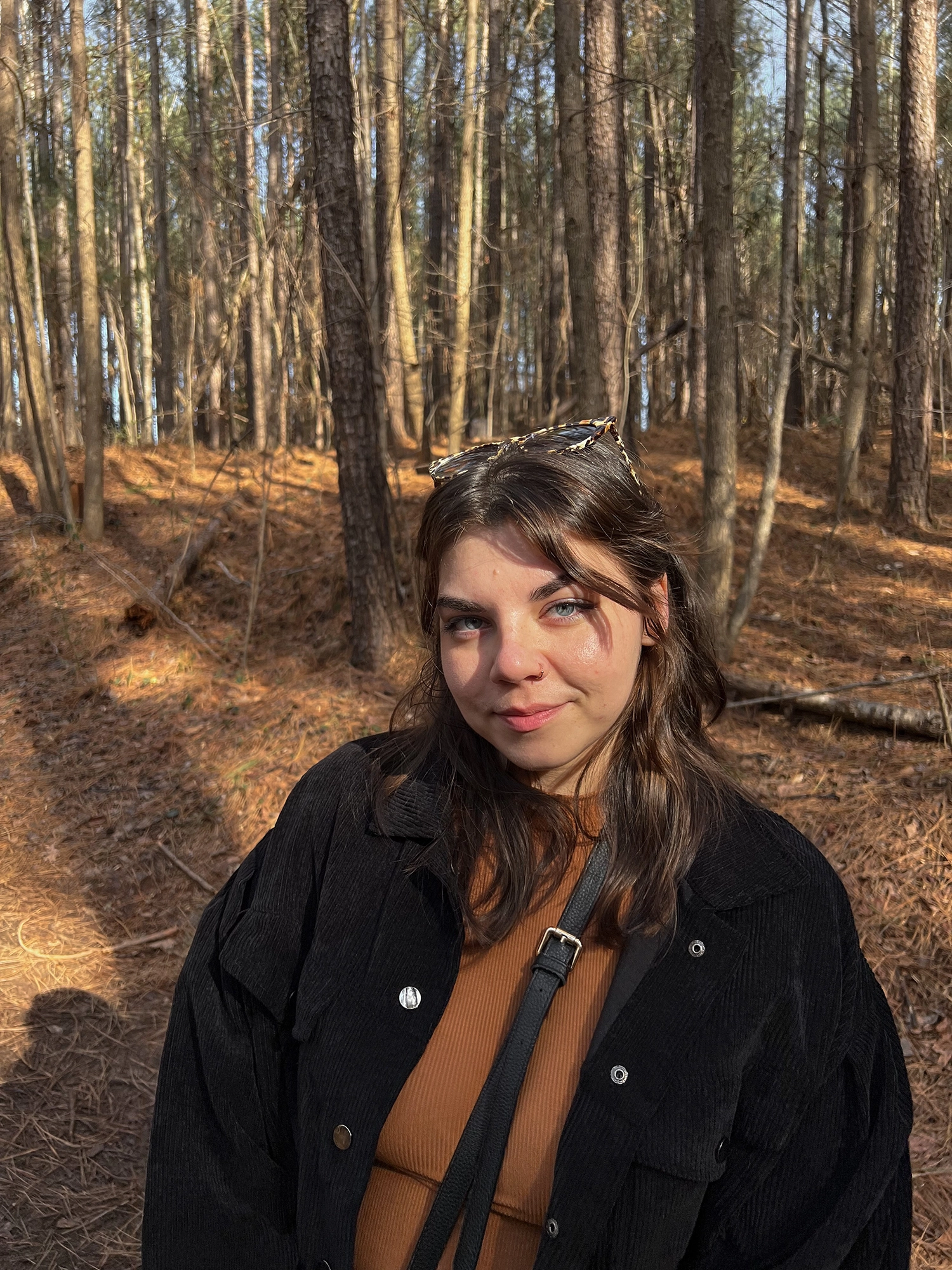 Young woman with brown hair and blue eyes standing in a forest with sunlight filtering through the trees.