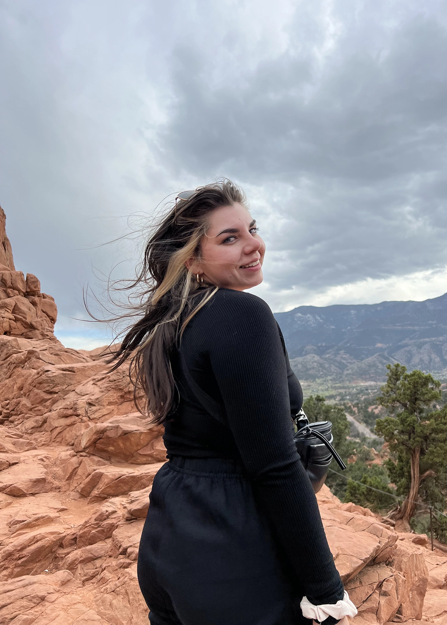 Smiling woman in black clothing standing on red rocky terrain with wind blowing her hair and mountains in the background under a cloudy sky.