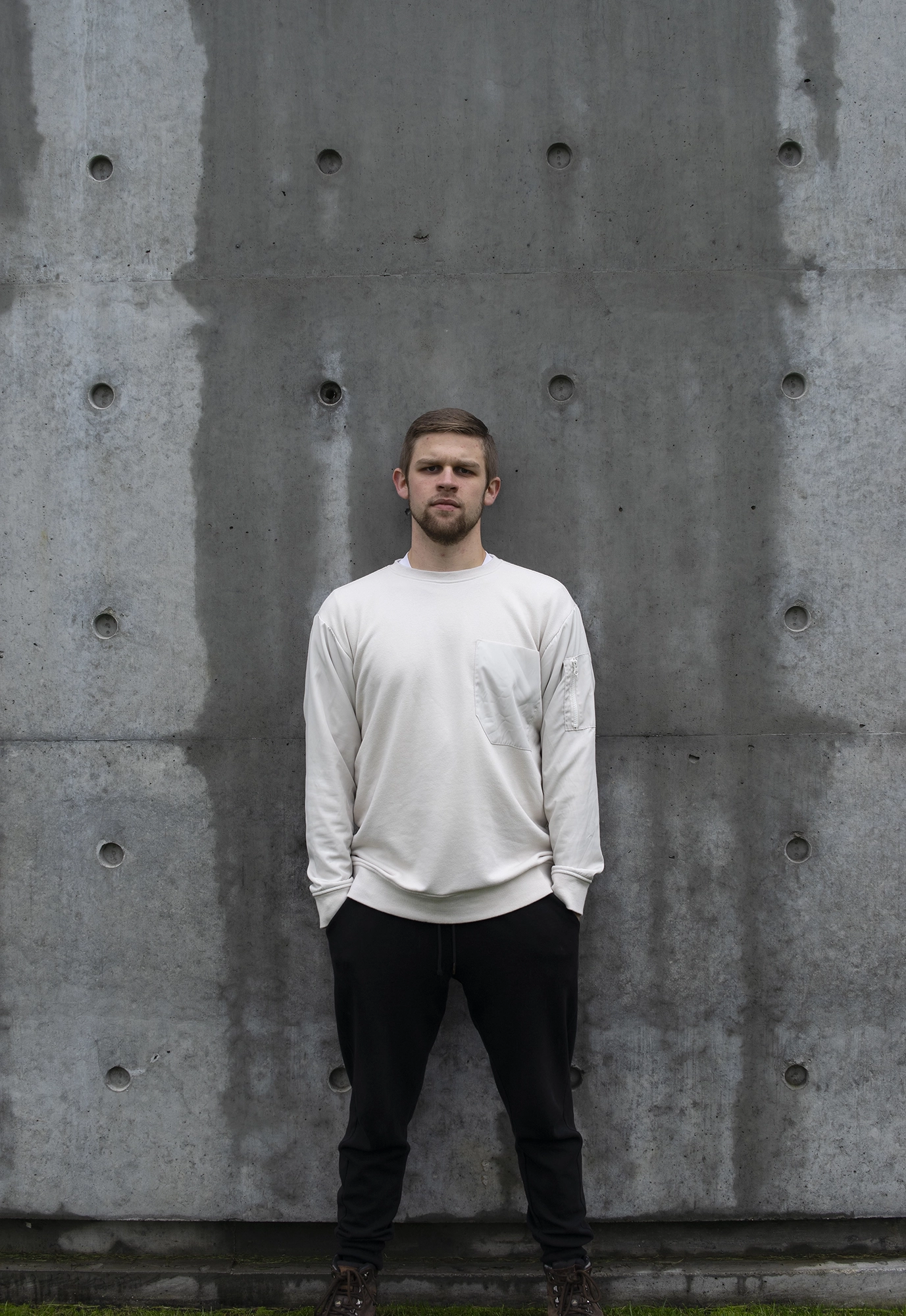 Stark image of a young man with short hair and a beard wearing a white sweatshirt standing with a serious expression against a textured concrete wall.
