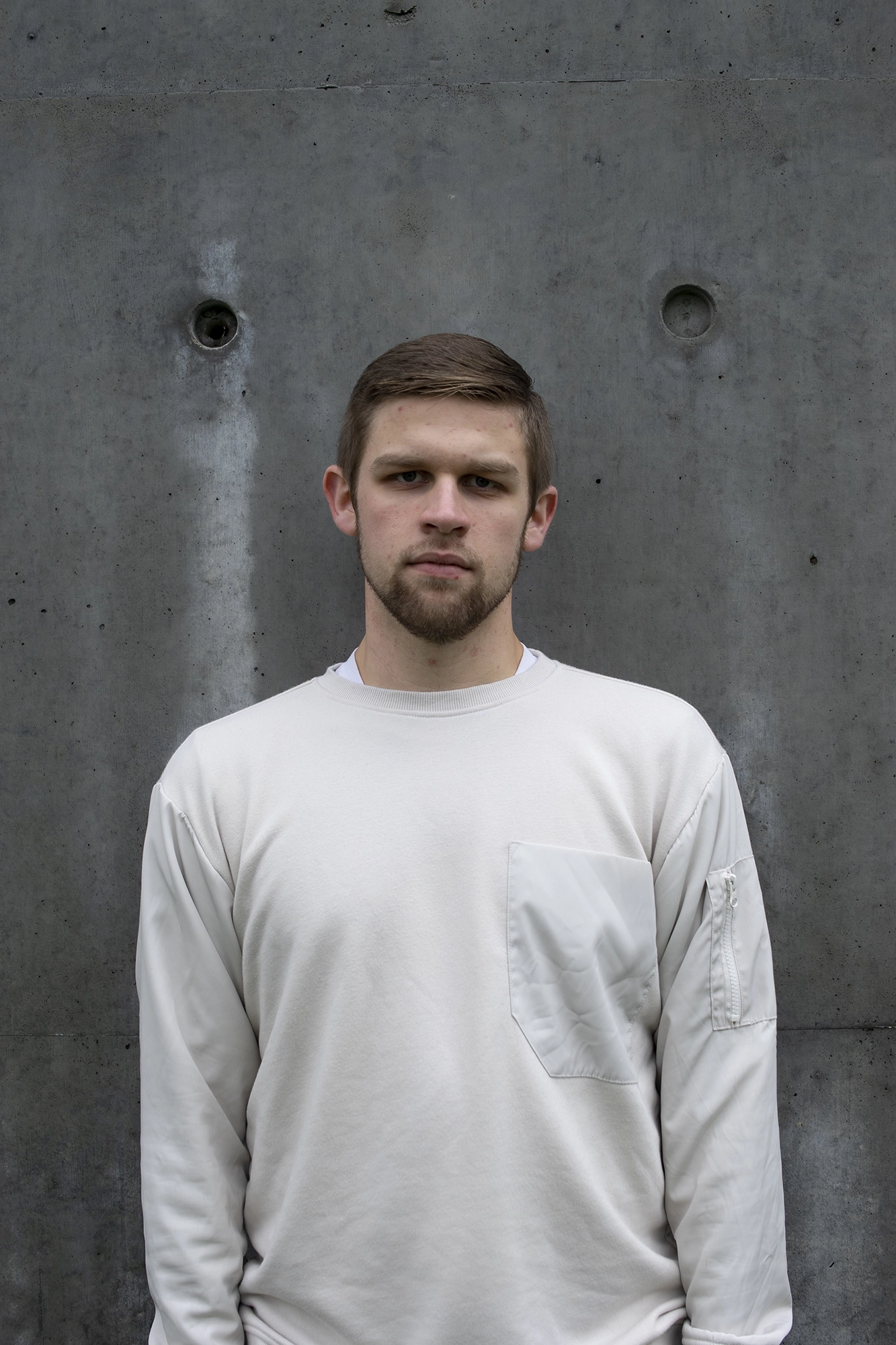 Stark image of a young man with short hair and a beard wearing a white sweatshirt standing with a serious expression against a textured concrete wall.