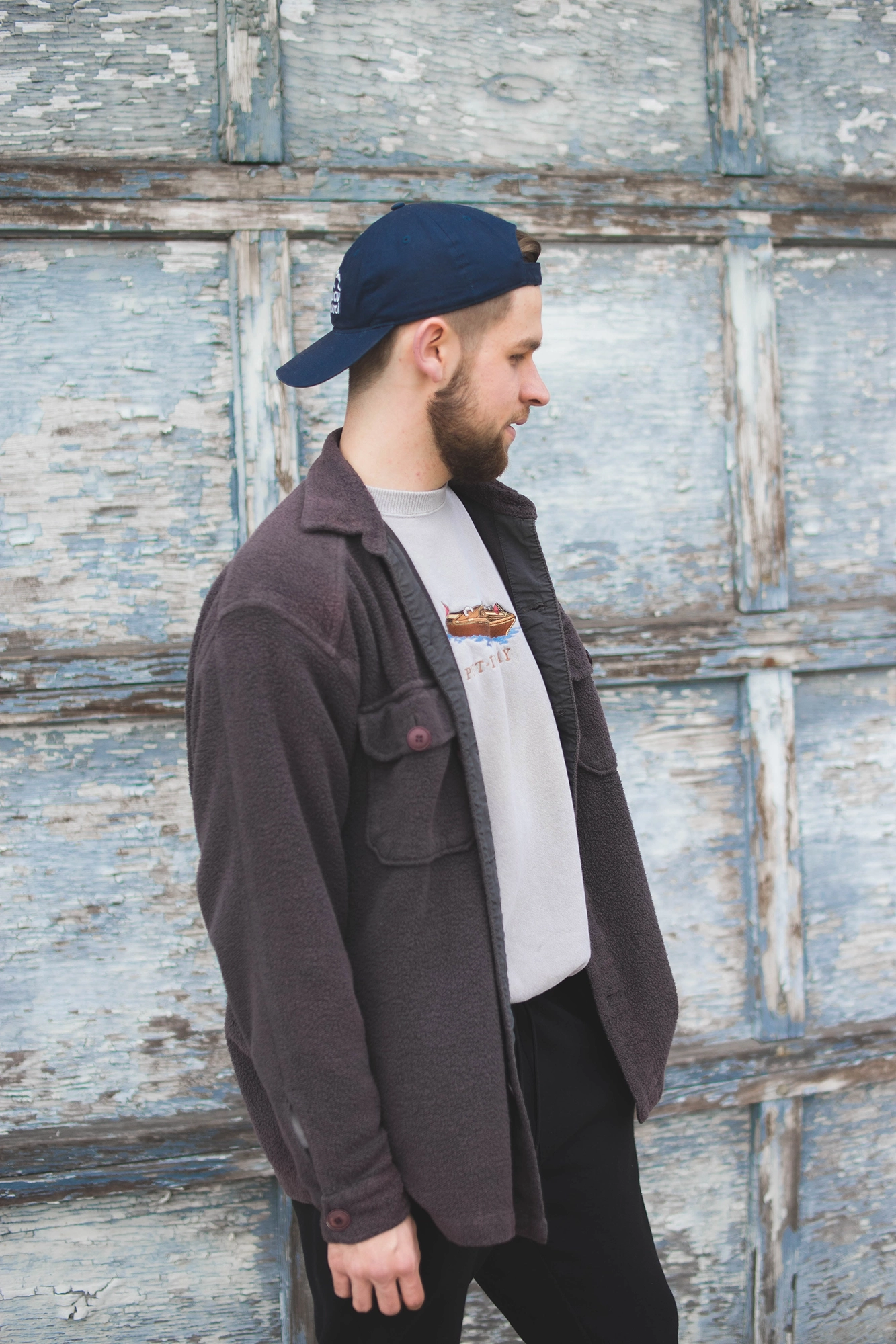 Side profile of a man wearing a navy blue backwards cap, brown jacket, and light gray sweatshirt standing against a weathered blue wooden wall.