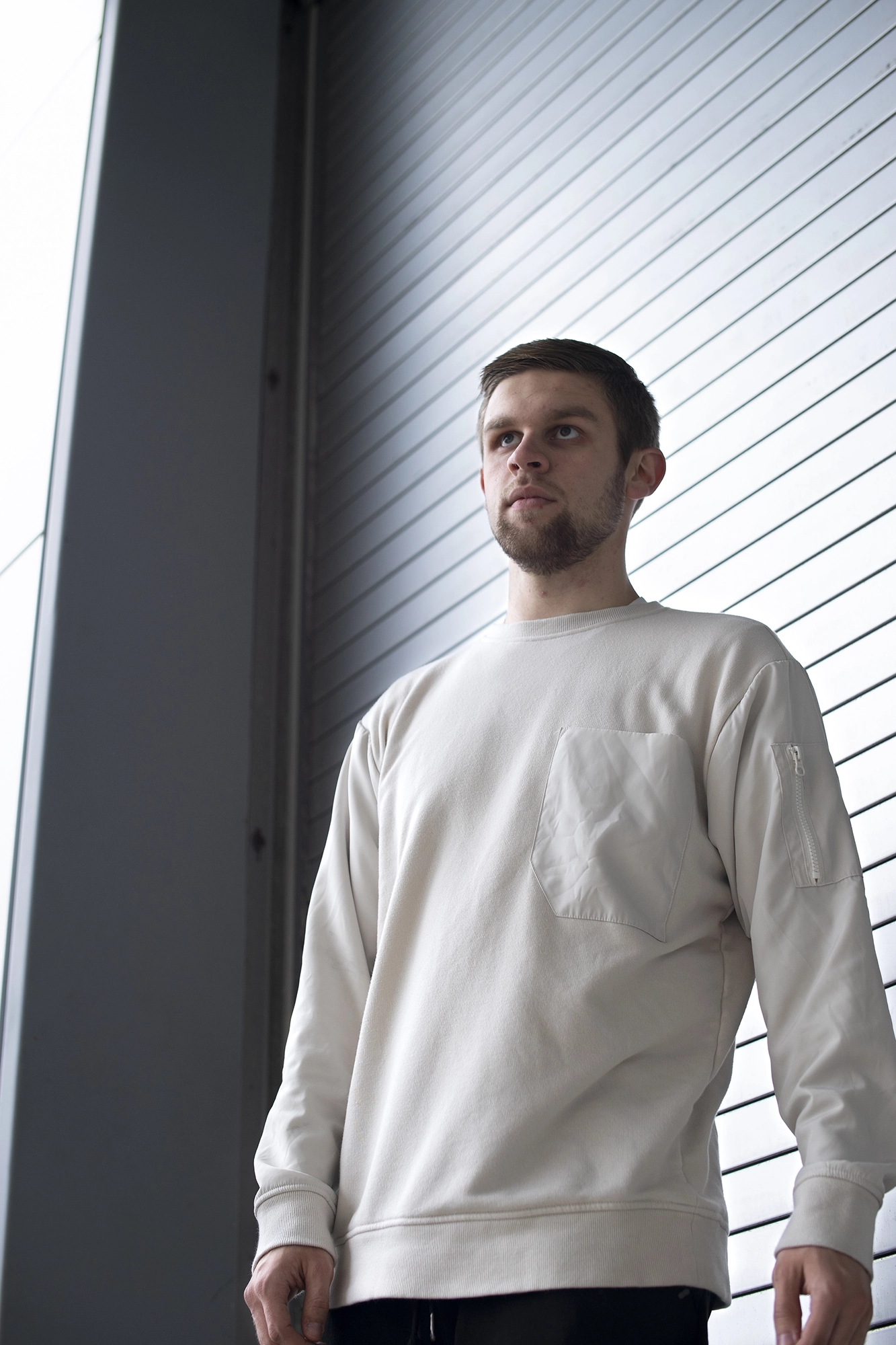 Young man with short hair, a beard, and a serious expression, wearing a white sweatshirt standing against a modern metallic wall with horizontal lines.
