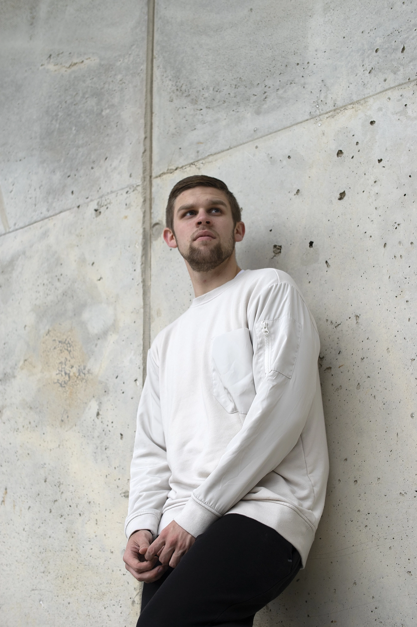 Young man wearing a white long-sleeve shirt and black pants leaning against a textured tan concrete wall, looking upward.