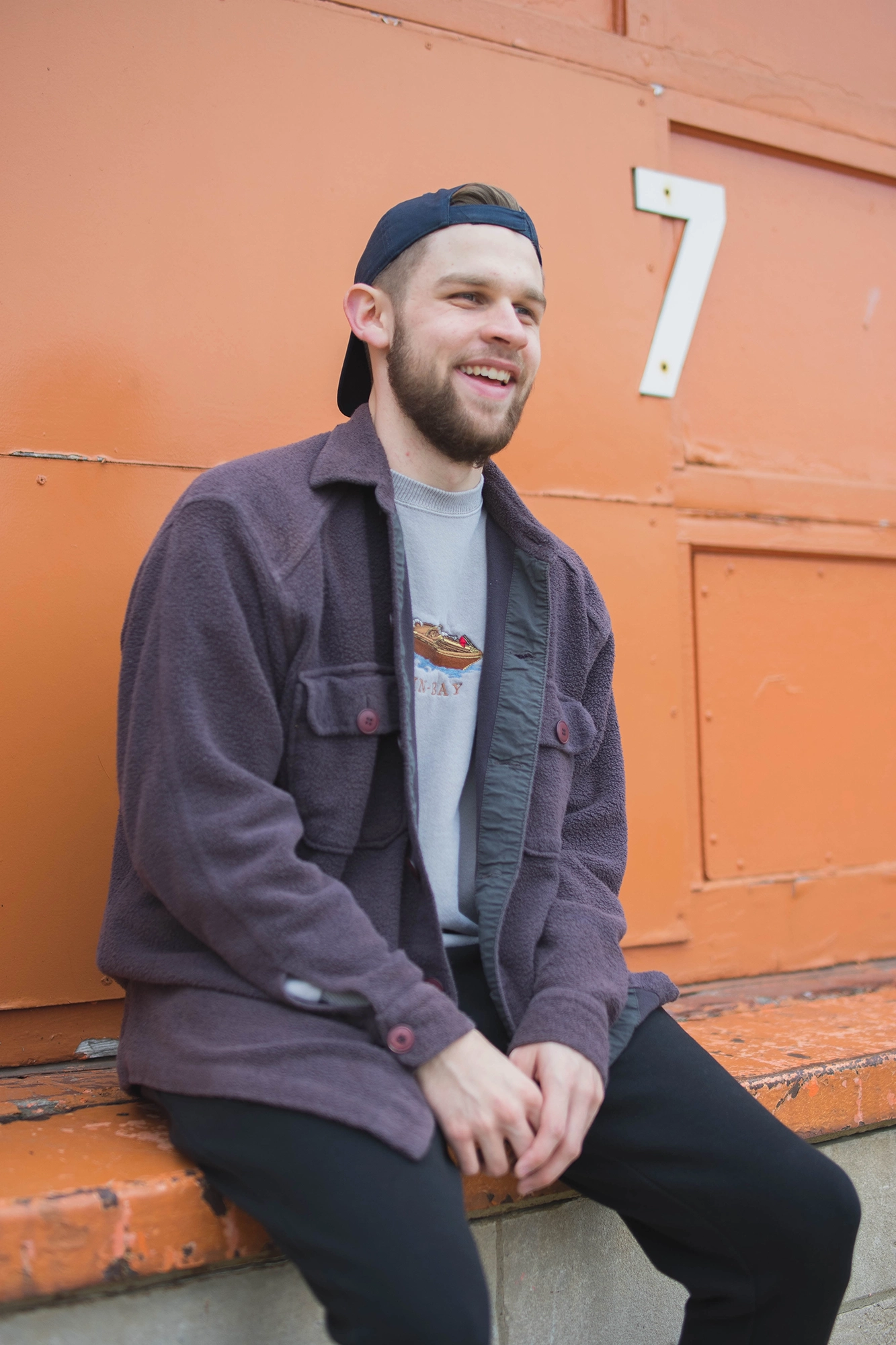 Smiling young man wearing a blue baseball cap backward, gray graphic sweatshirt, and purple jacket sitting against an orange wall with a white number 7.