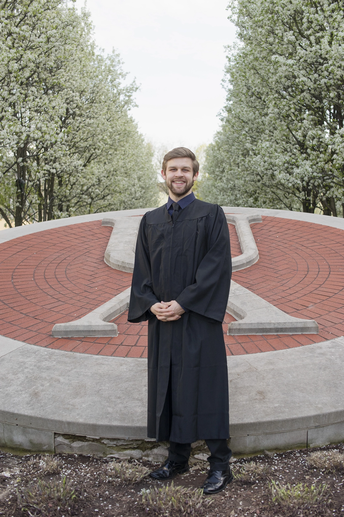 Smiling young man in graduation gown standing in front of a large red and concrete university emblem surrounded by blossoming trees.