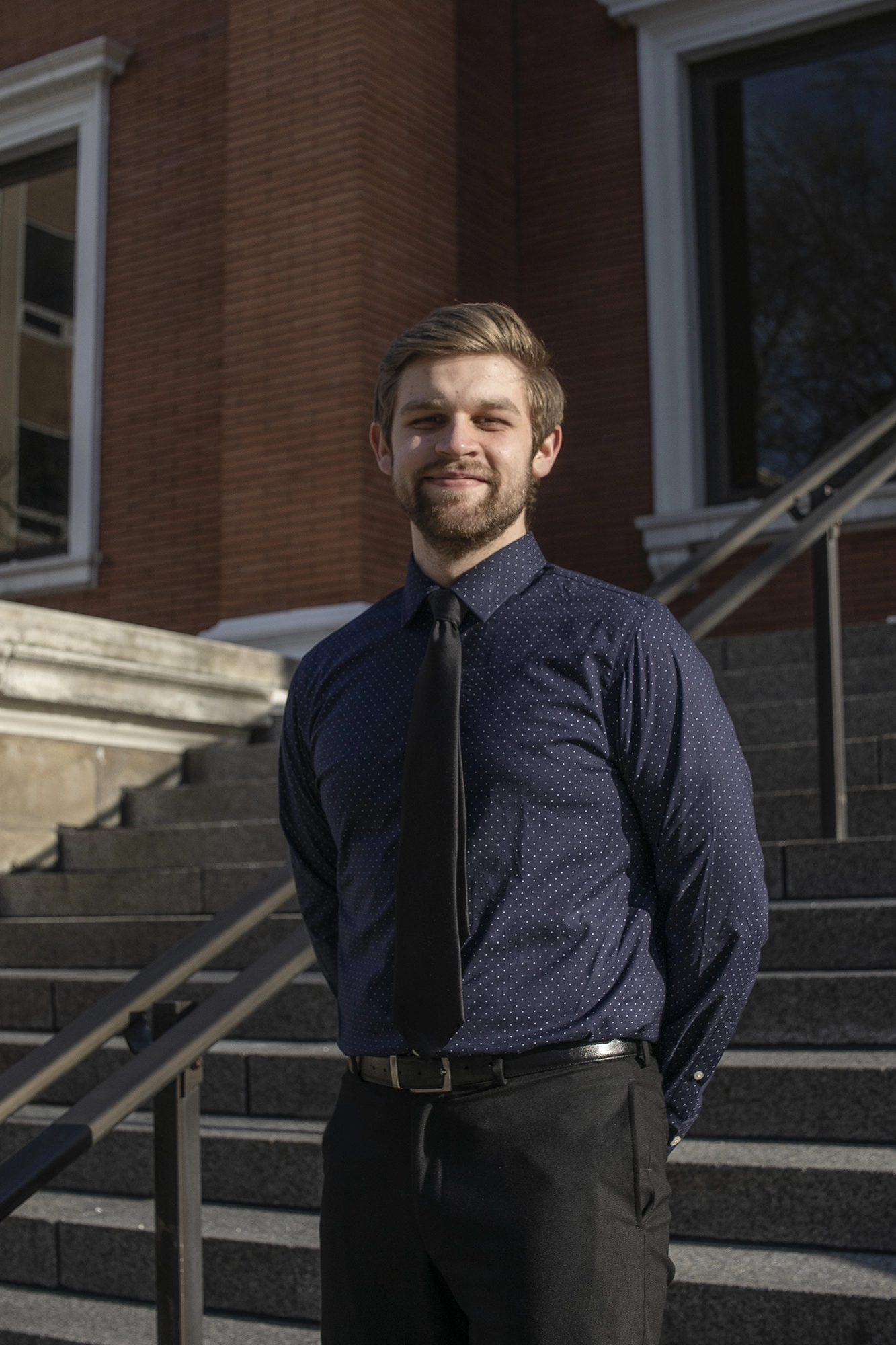 Young man in a navy blue polka dot shirt and black tie standing outdoors in front of stone steps and a brick building.
