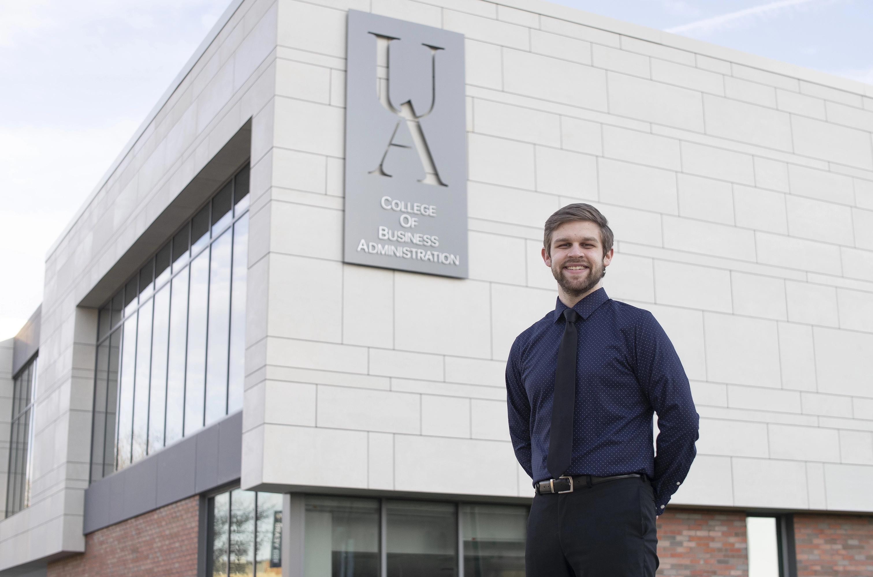 Smiling young man in blue polka dot shirt and black tie standing in front of UA College of Business Administration building.