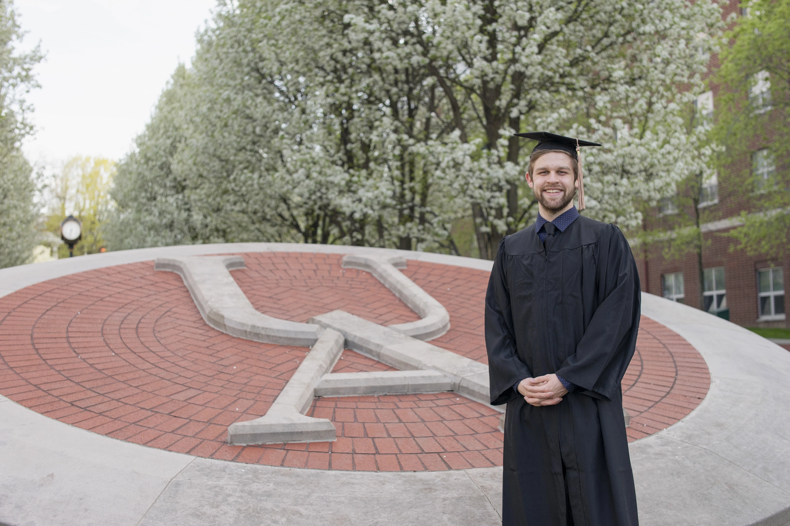 Young male graduate in black cap and gown standing and smiling in front of a large circular brick and concrete university emblem outdoors with blooming trees in the background.