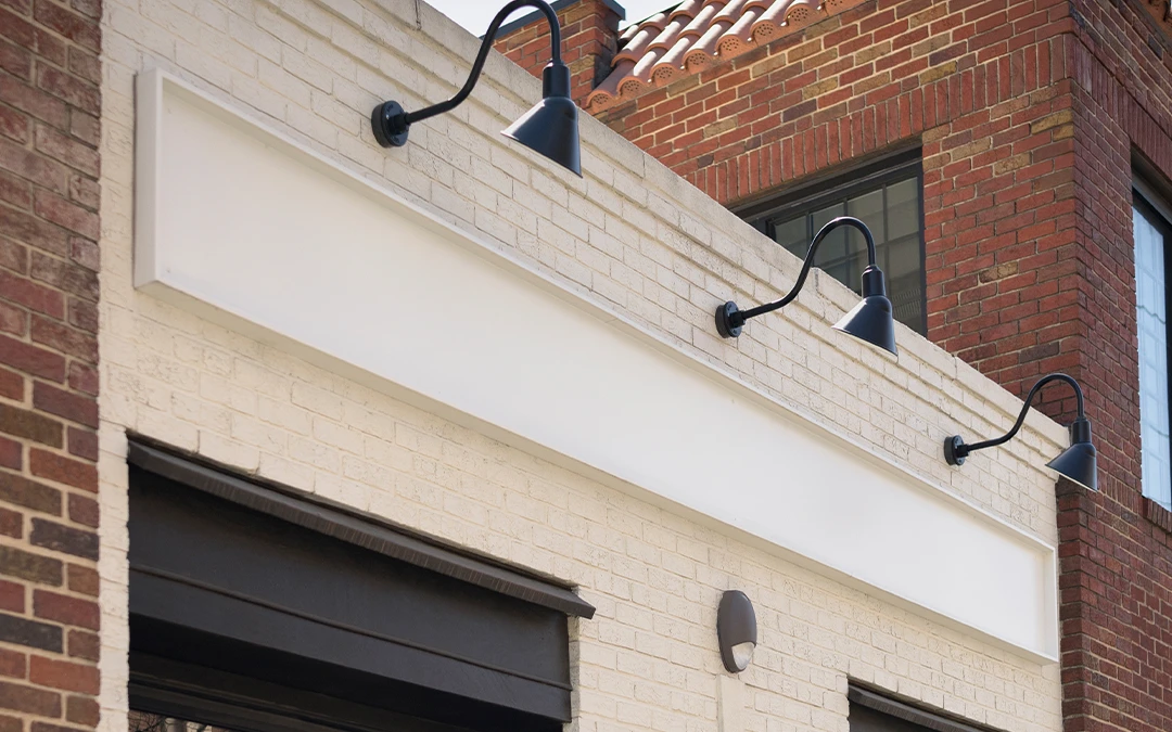 White brick storefront with black framed windows and a rectangular sign under three black gooseneck lights.