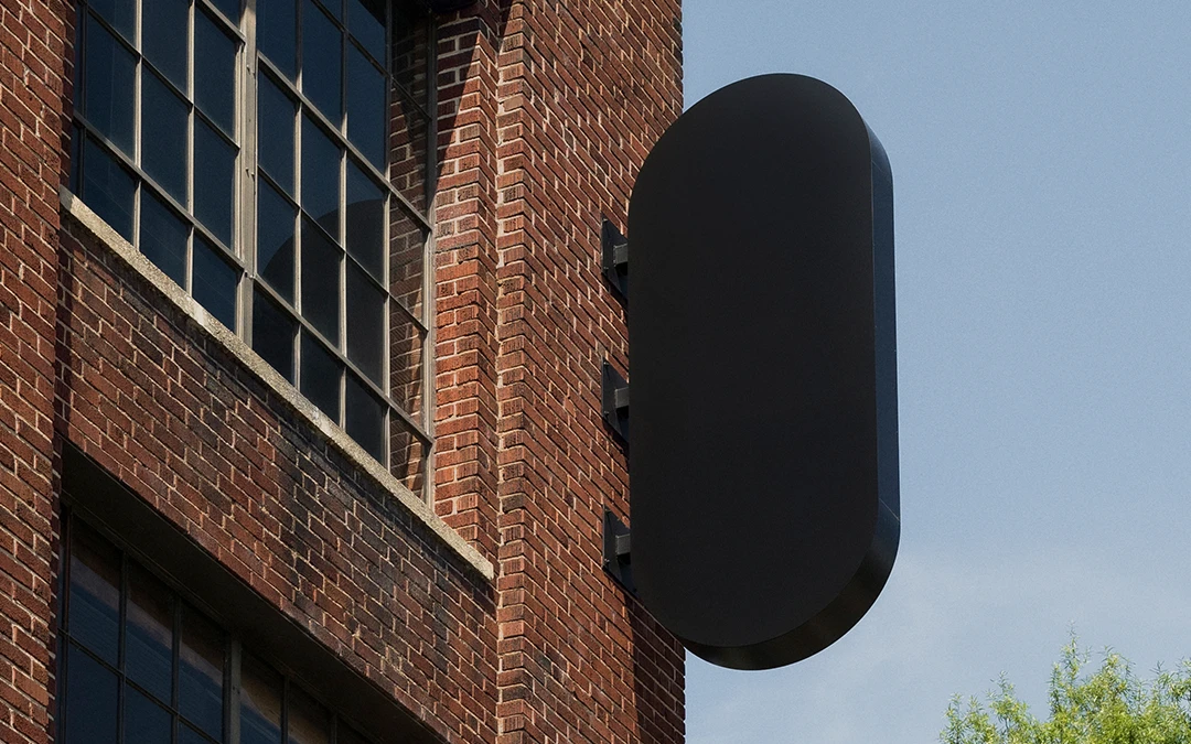 Black pill-shaped signage mounted on a brick building under a clear blue sky with green trees nearby.