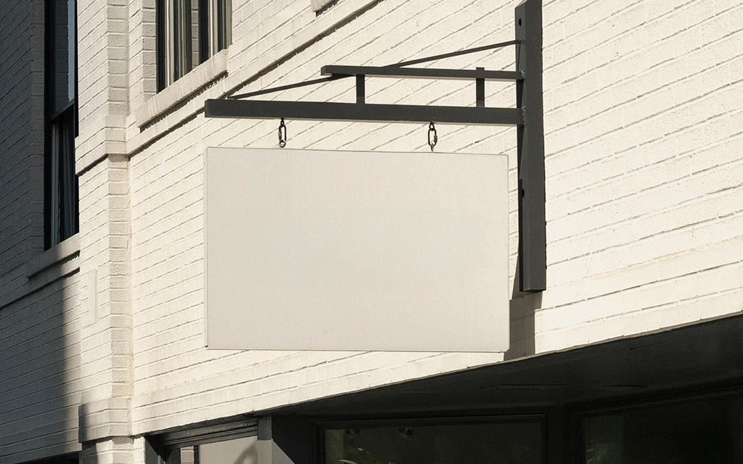 Off-white hanging sign mounted on a light brick building facade next to green tree branches.