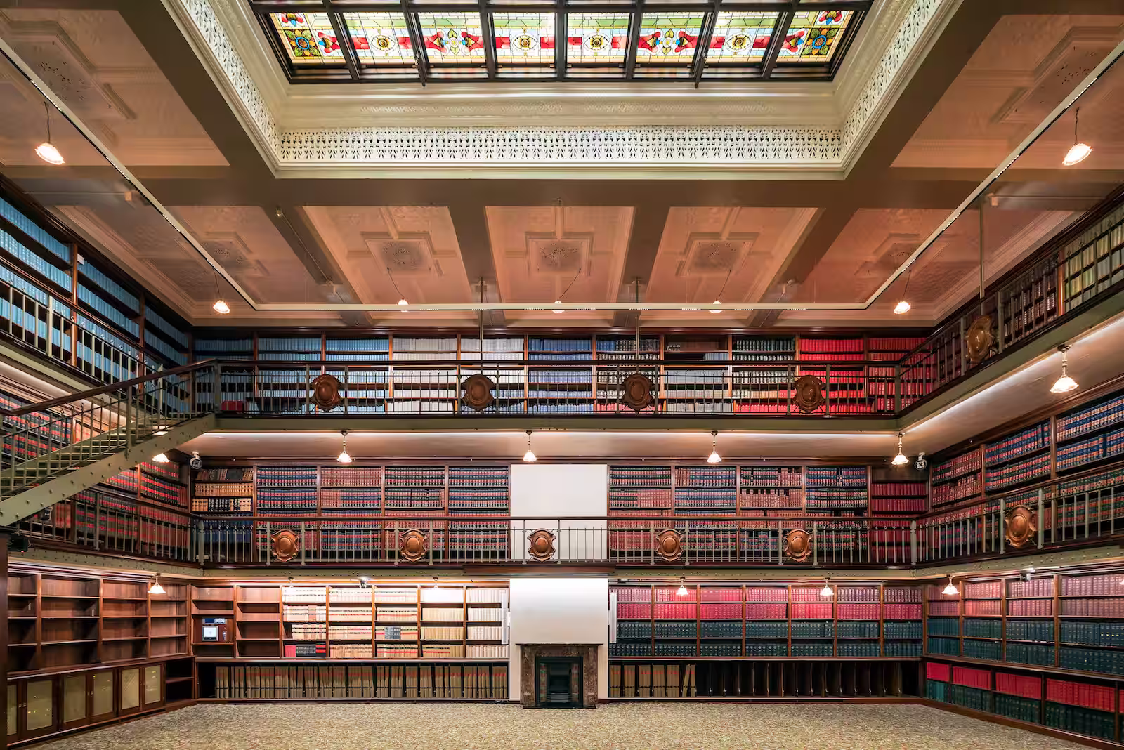 Interior of a large, historic library with multiple levels of wooden bookshelves filled with books and a stained glass skylight ceiling.