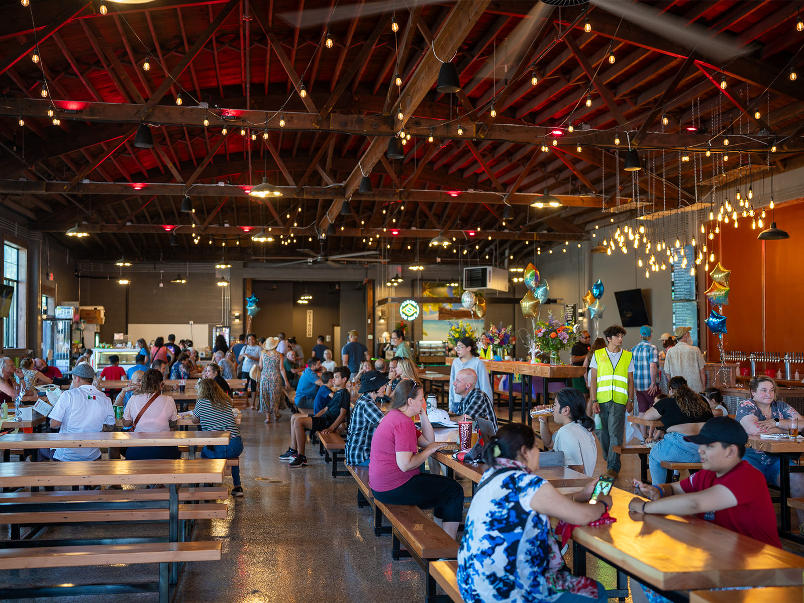 Diners at long communal wood tables beneath the exposed wood truss ceiling and string lights inside Hillsboro Downtown Station.