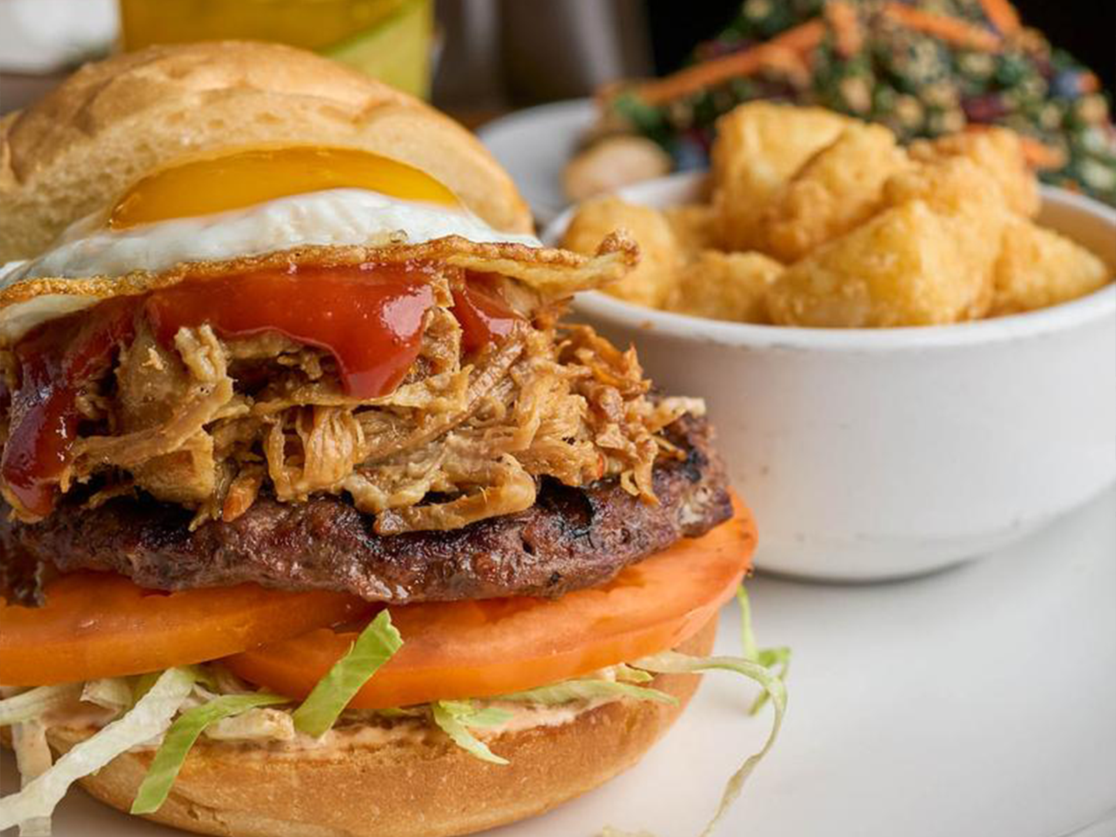 A loaded burger topped with pulled pork and a fried egg, served with crispy tots and a kale salad at BRIX Tavern