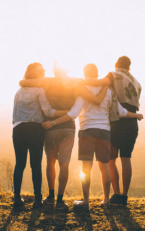 Four people standing side by side with arms around each other facing a sunset over a grassy landscape.