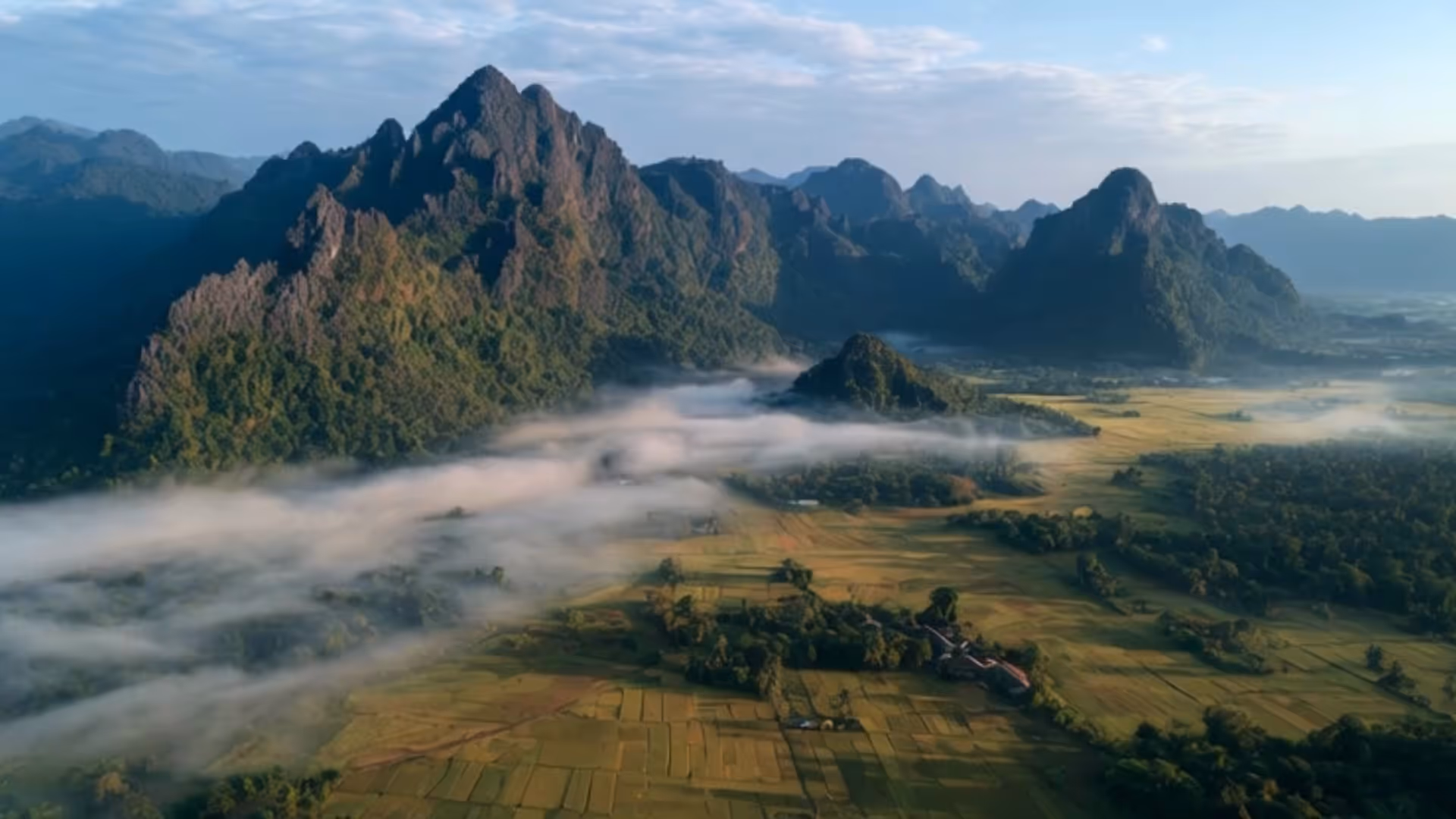 Mountain range with green vegetation and mist floating over a patchwork of fields under a blue sky.