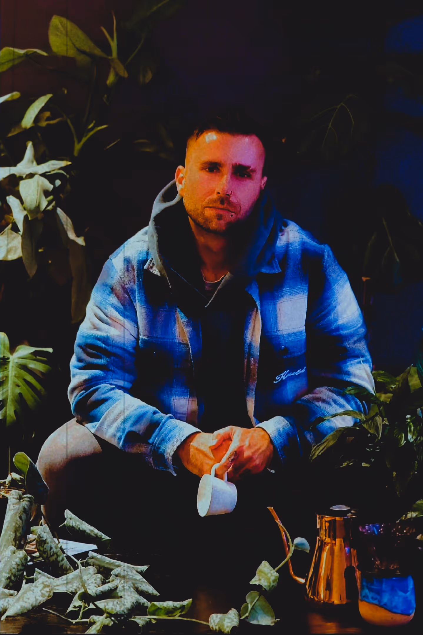 Logan. the founder of Logies Lab, in a blue plaid jacket holding a white cup, surrounded by green plants and a copper kettle on a table in dim lighting.