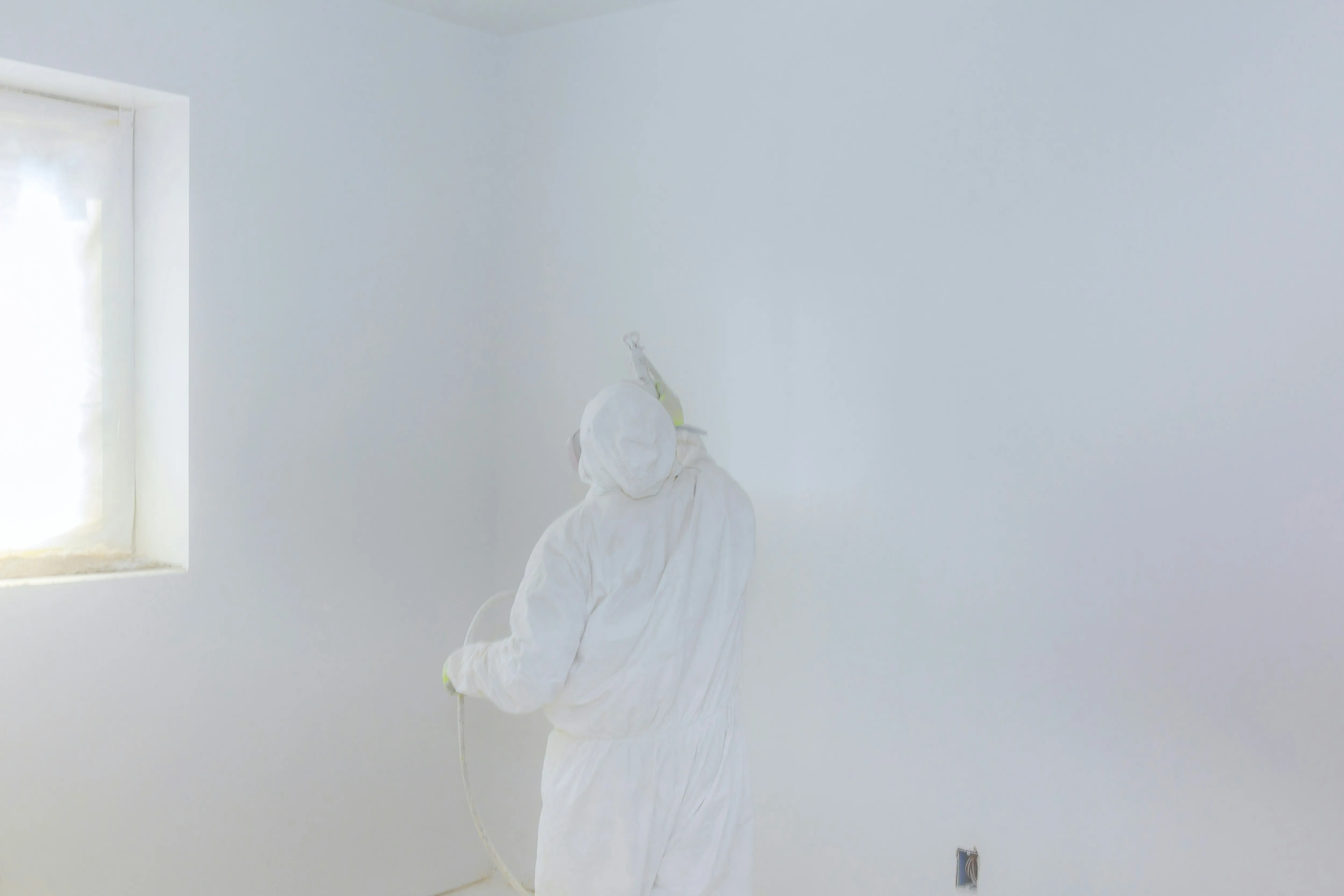 Stucadoor in beschermende kleding brengt witte verf aan met sausspuiten op stucwerk bij raam
