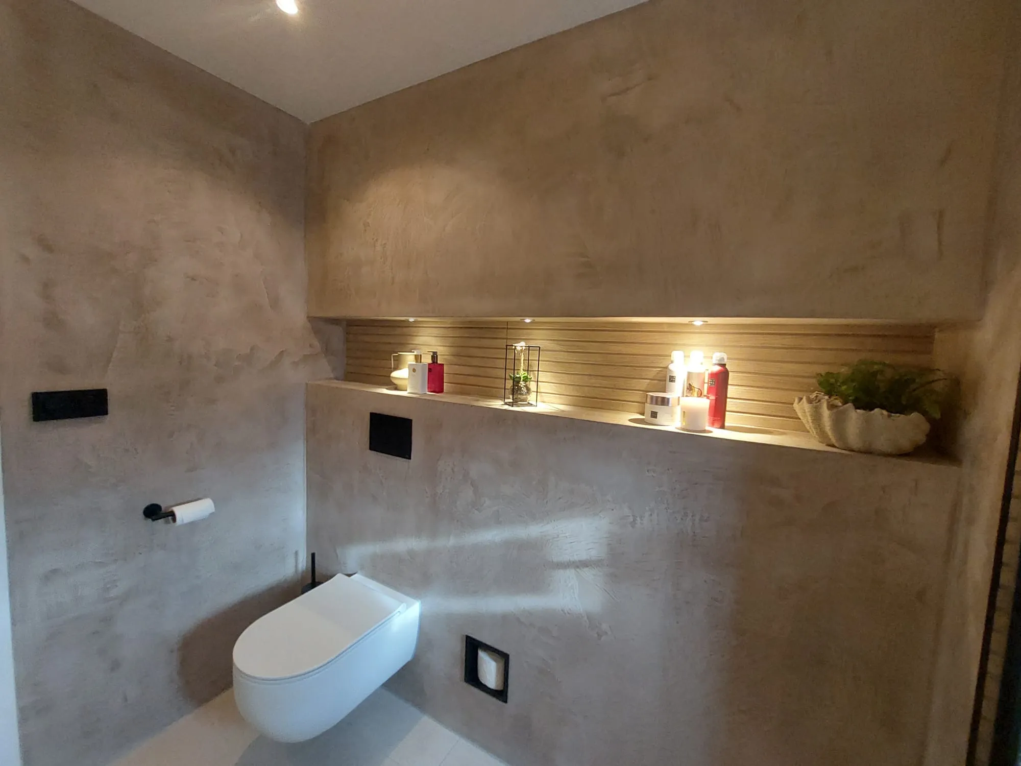 Modern toilet met beige sierpleister gestructureerd stucwerk, zwevende WC en ingebouwde plank onder warme verlichting
